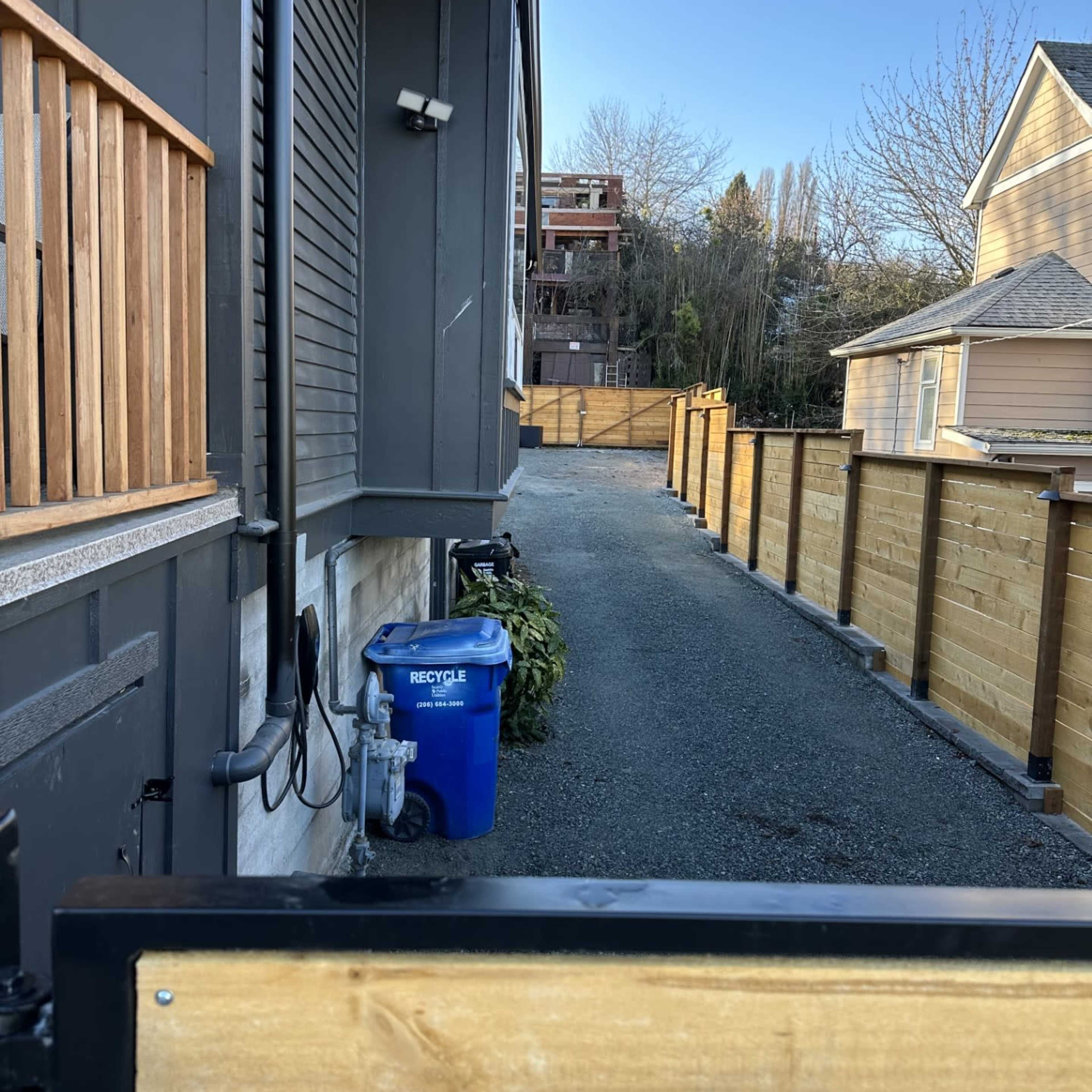 A narrow gravel path runs between two houses, with a blue recycling bin and wooden fences lining either side.