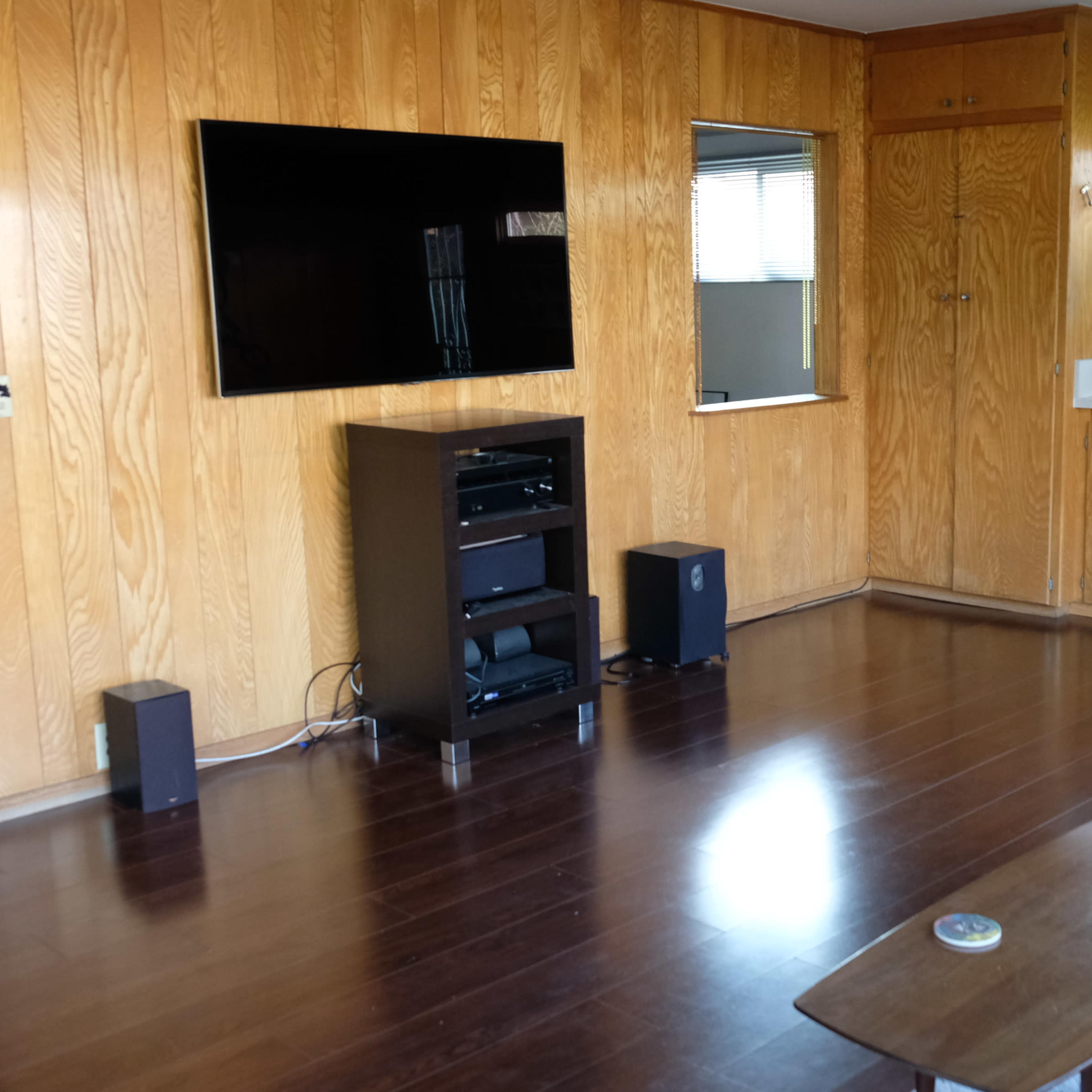 A living room with wooden paneling, a wall-mounted TV, a black entertainment unit, and a small coffee table on a patterned rug.