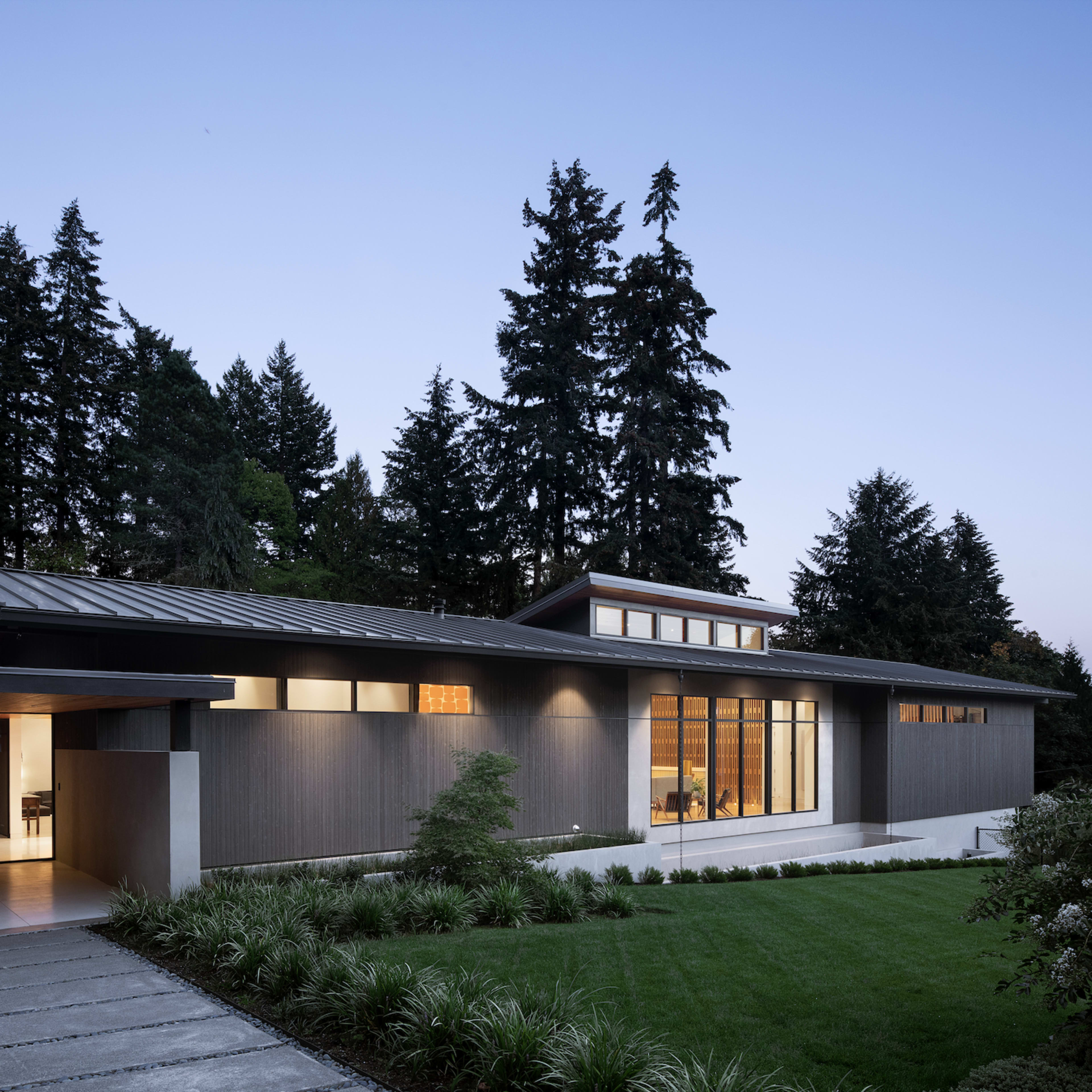 A modern house with a flat roof and large windows is situated amidst tall trees, featuring a landscaped front yard with greenery.