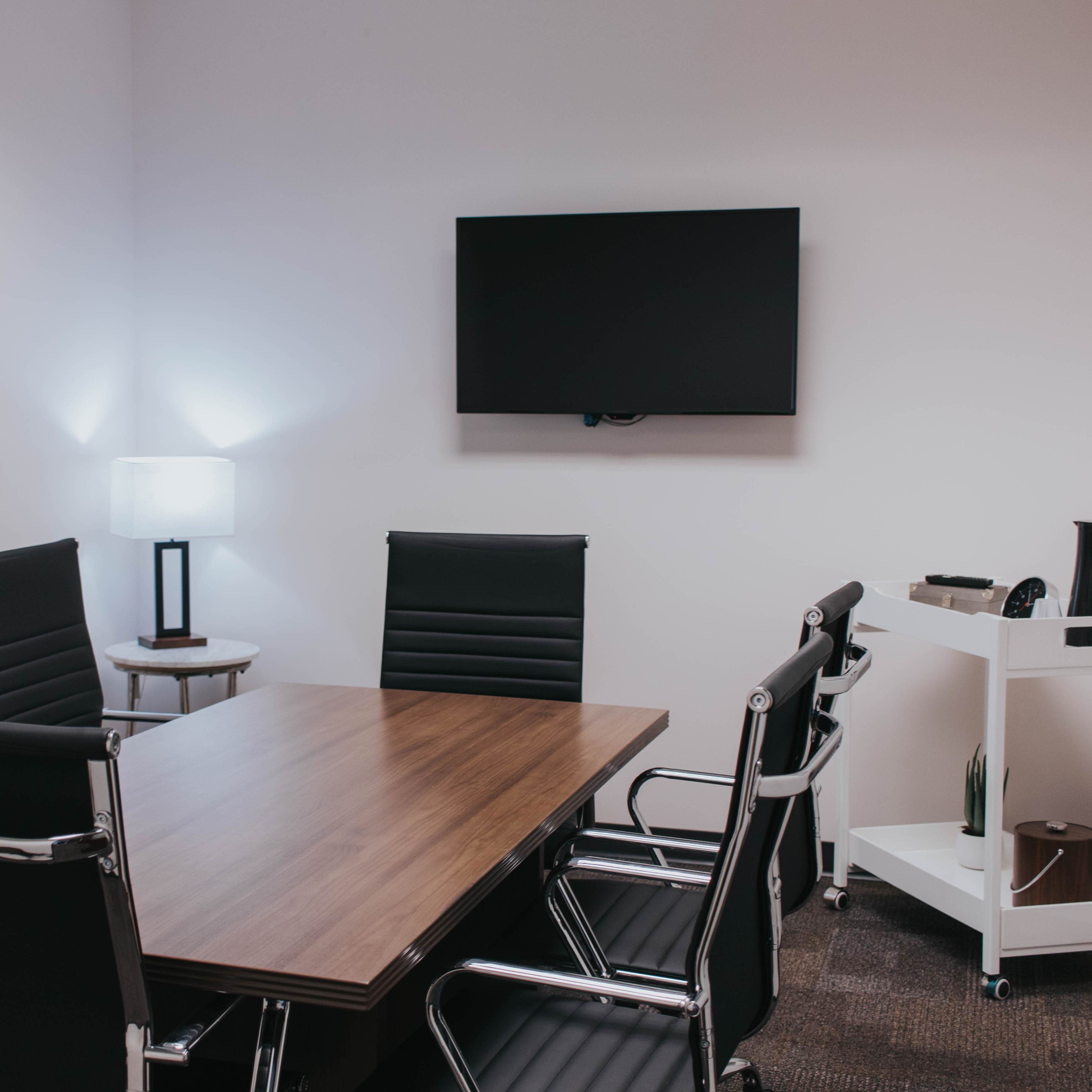 A modern conference room features a wooden table surrounded by sleek black chairs, a wall-mounted TV, a lamp, and a rolling cart with a coffee pot.