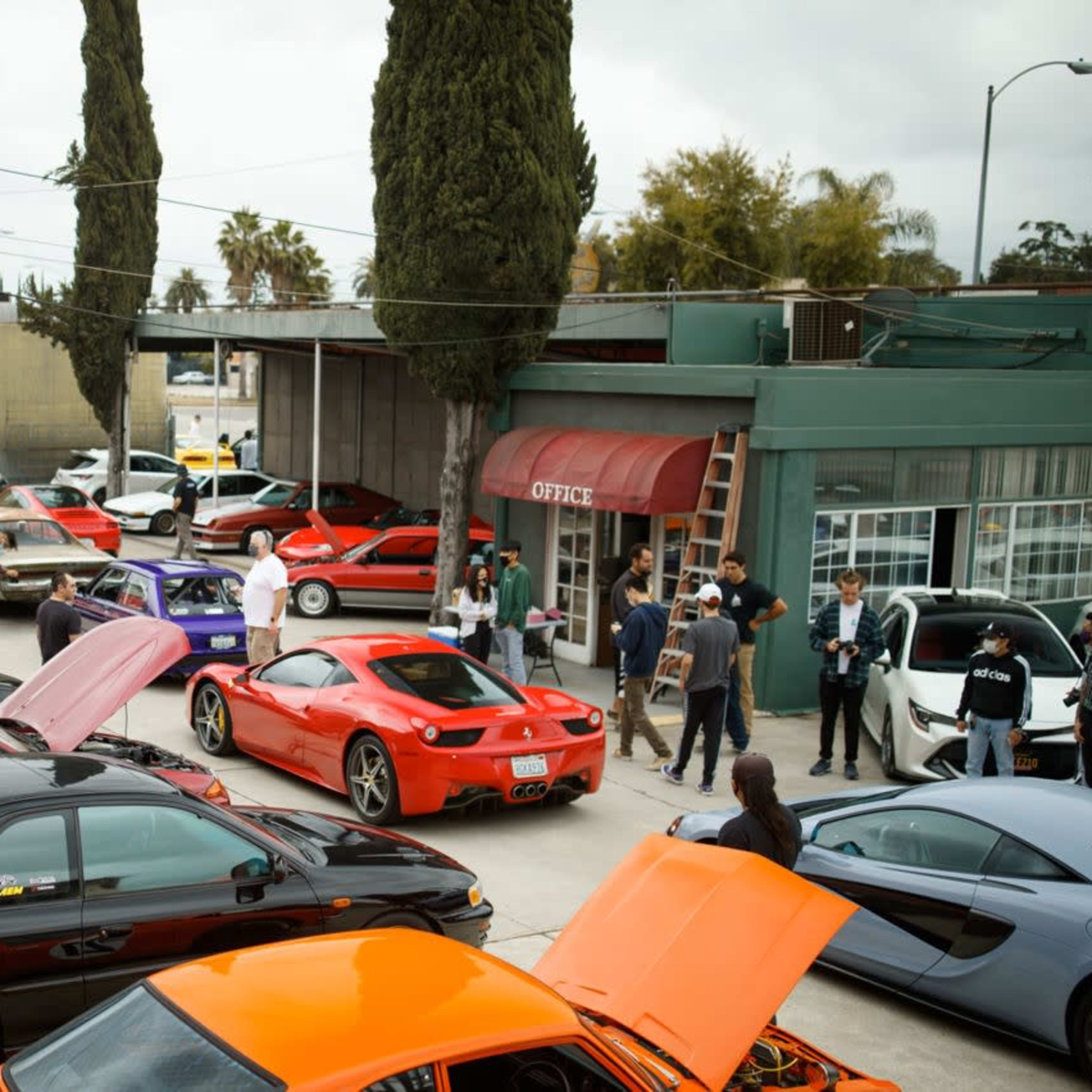 A gathering of various cars, including sports cars and modified vehicles, takes place in a parking lot surrounded by trees and buildings.