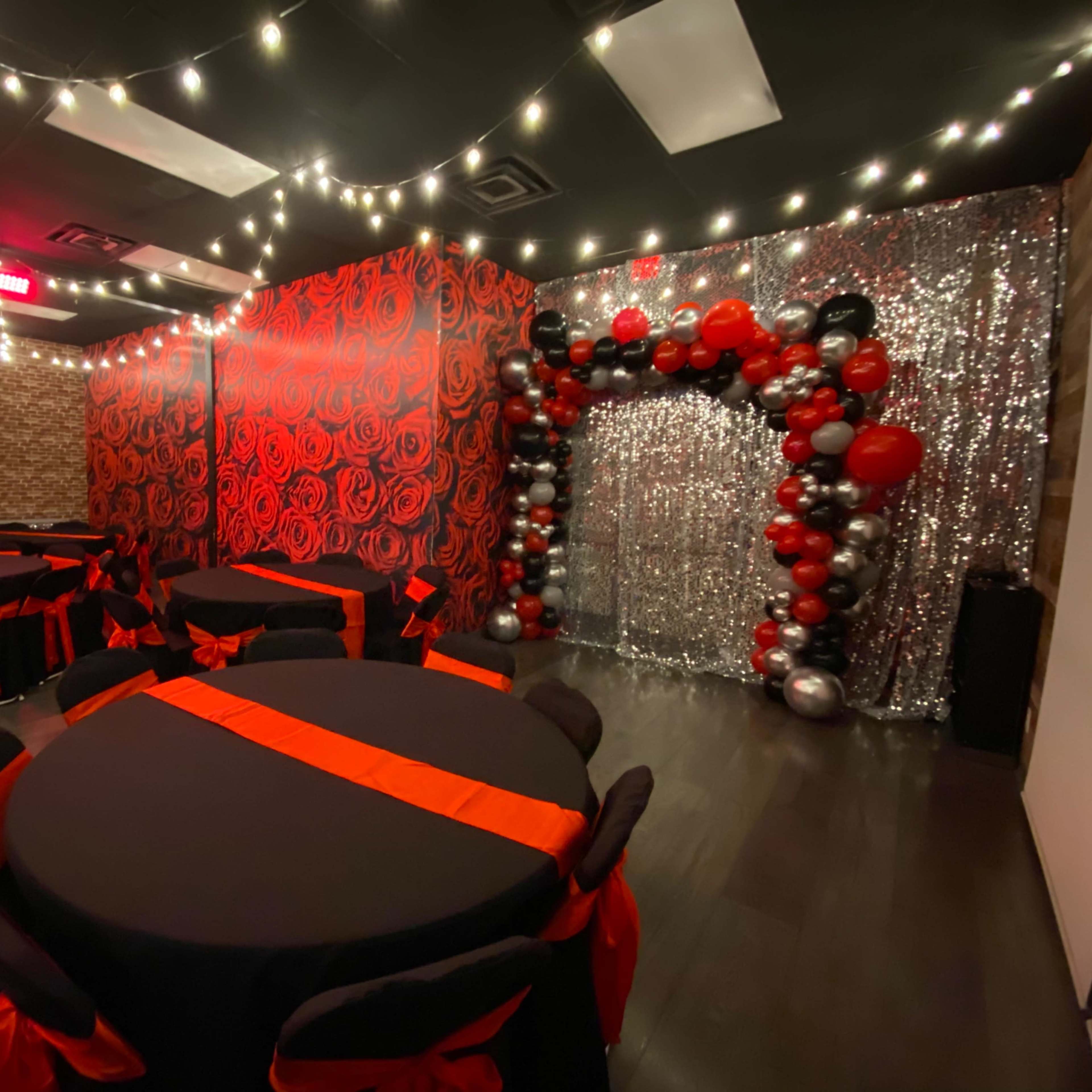 A decorated event space with black tables featuring red sashes, a wall adorned with red roses, and an arch made of silver and black balloons against a shimmering backdrop.