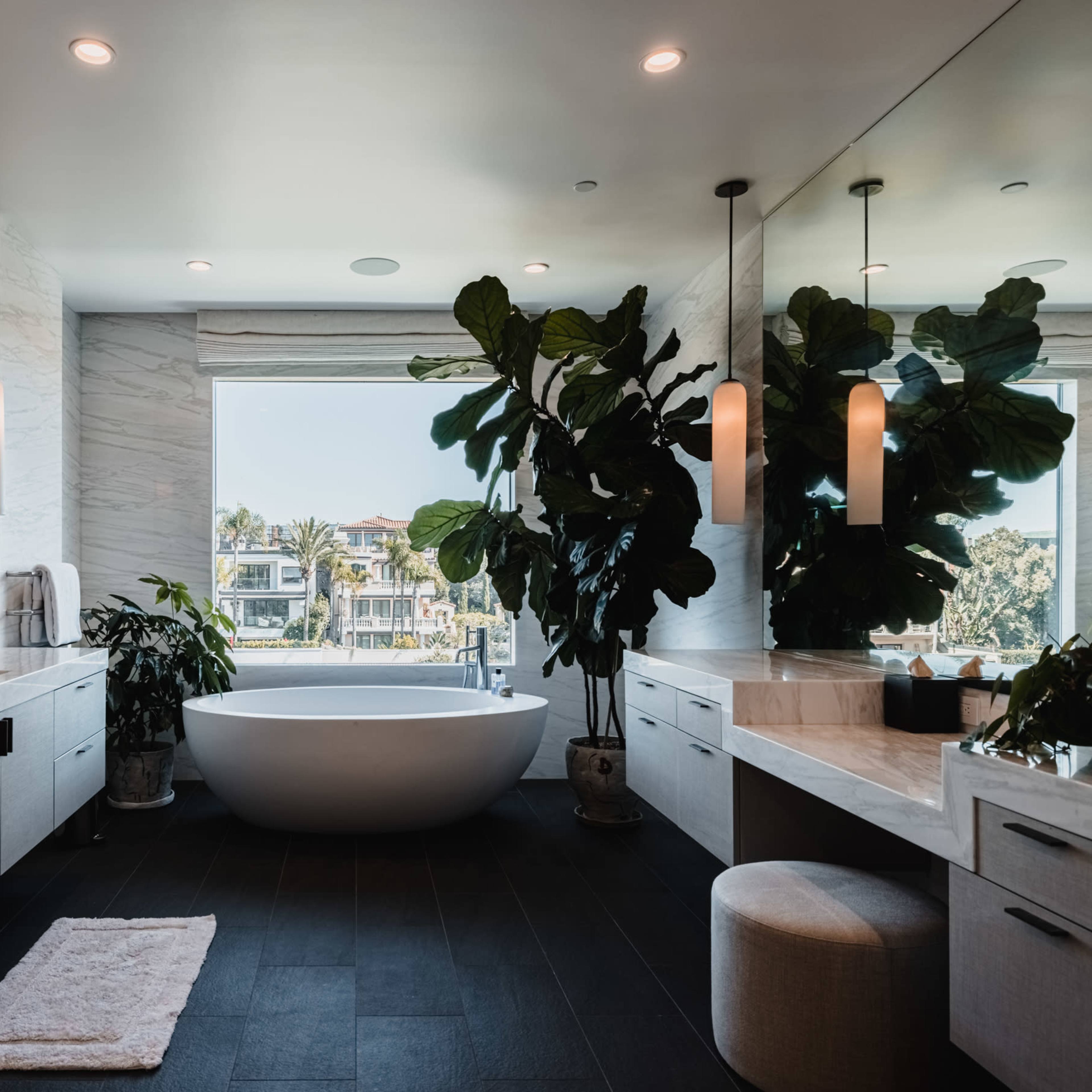 A modern bathroom features a freestanding bathtub, large windows with a view, and greenery in elegant pots.