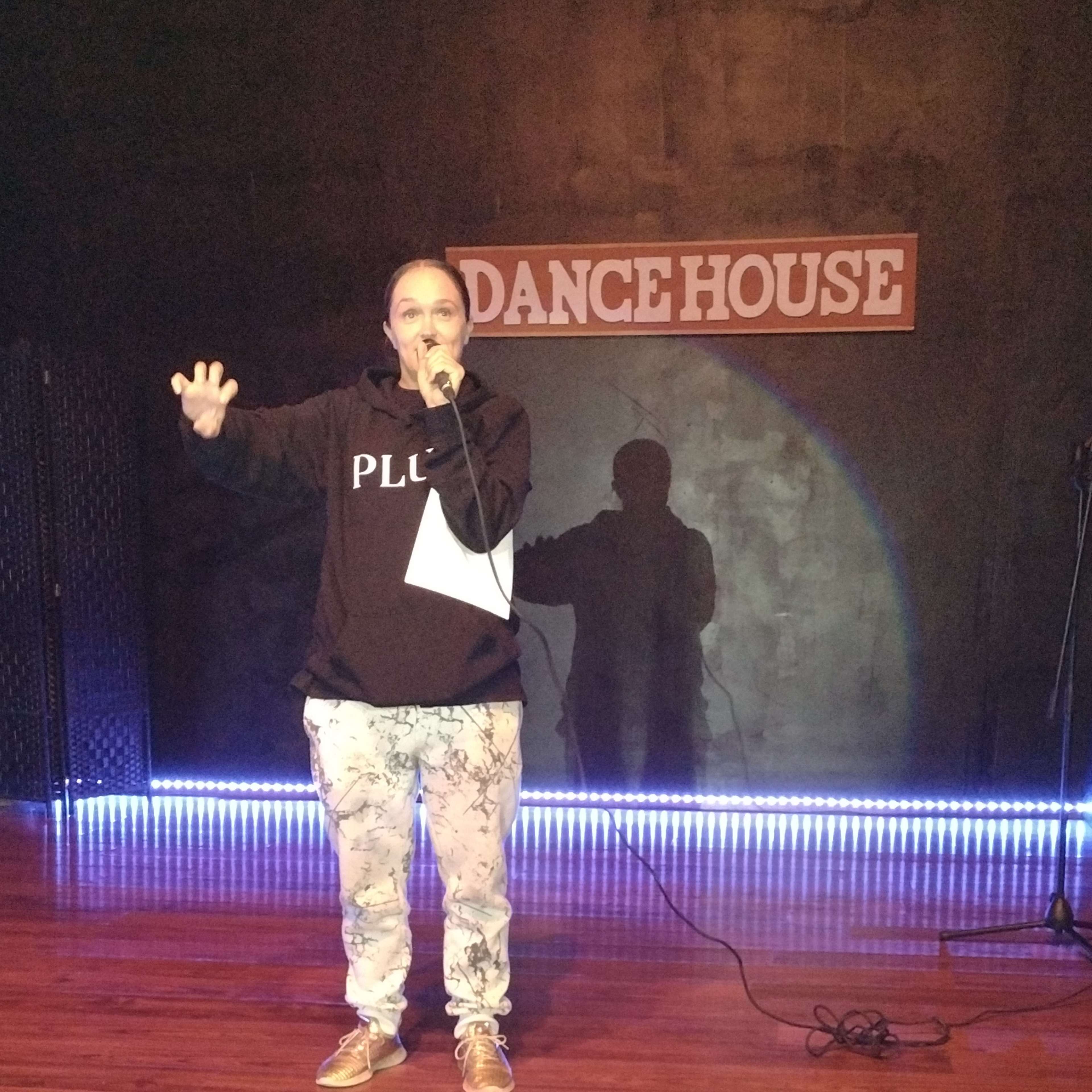 A person stands on a wooden stage in a performance space, holding a microphone and facing an audience with a shadow cast on the wall behind them.