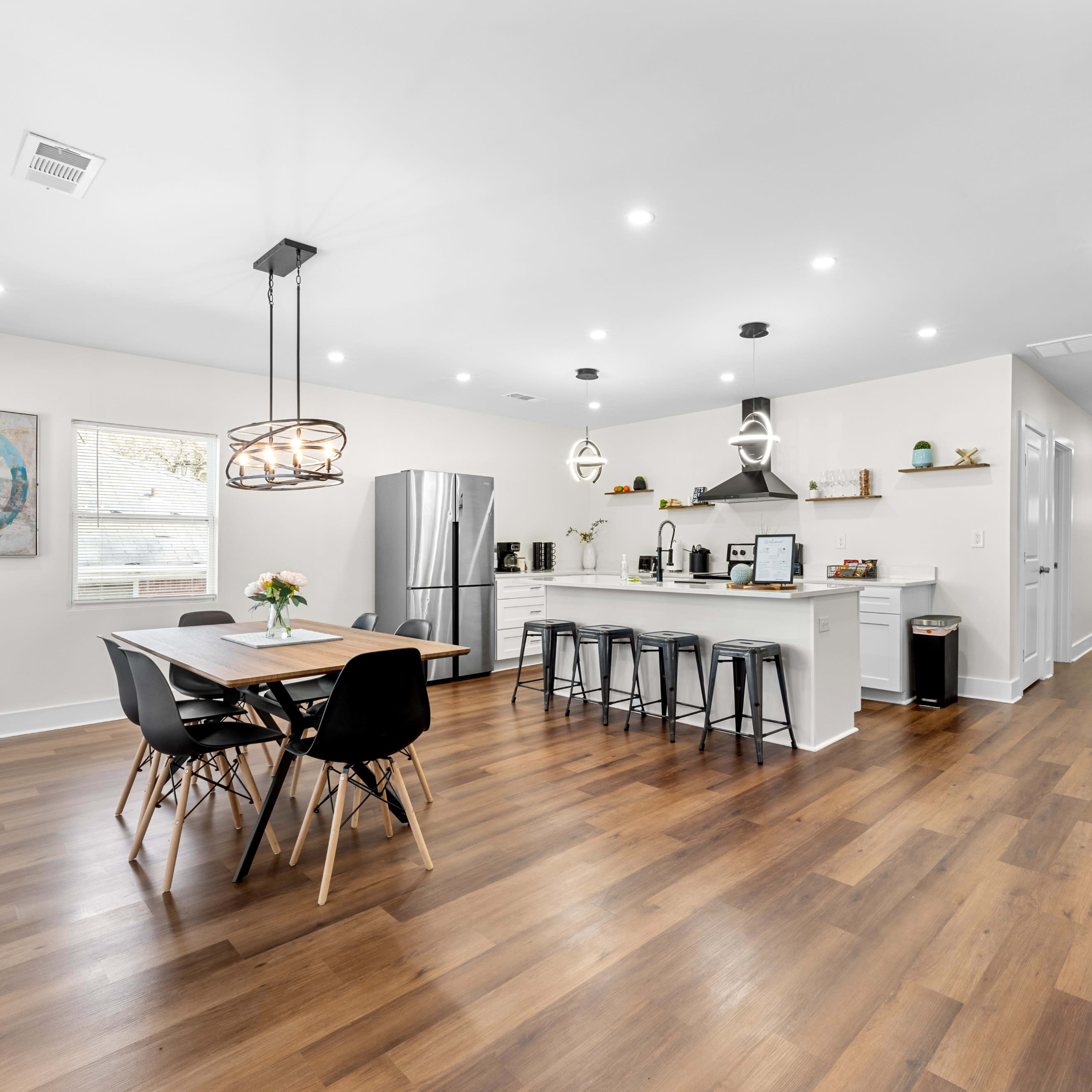 A modern kitchen and dining area features a long wooden table surrounded by black chairs, with a sleek kitchen equipped with stainless steel appliances and a minimalist design.