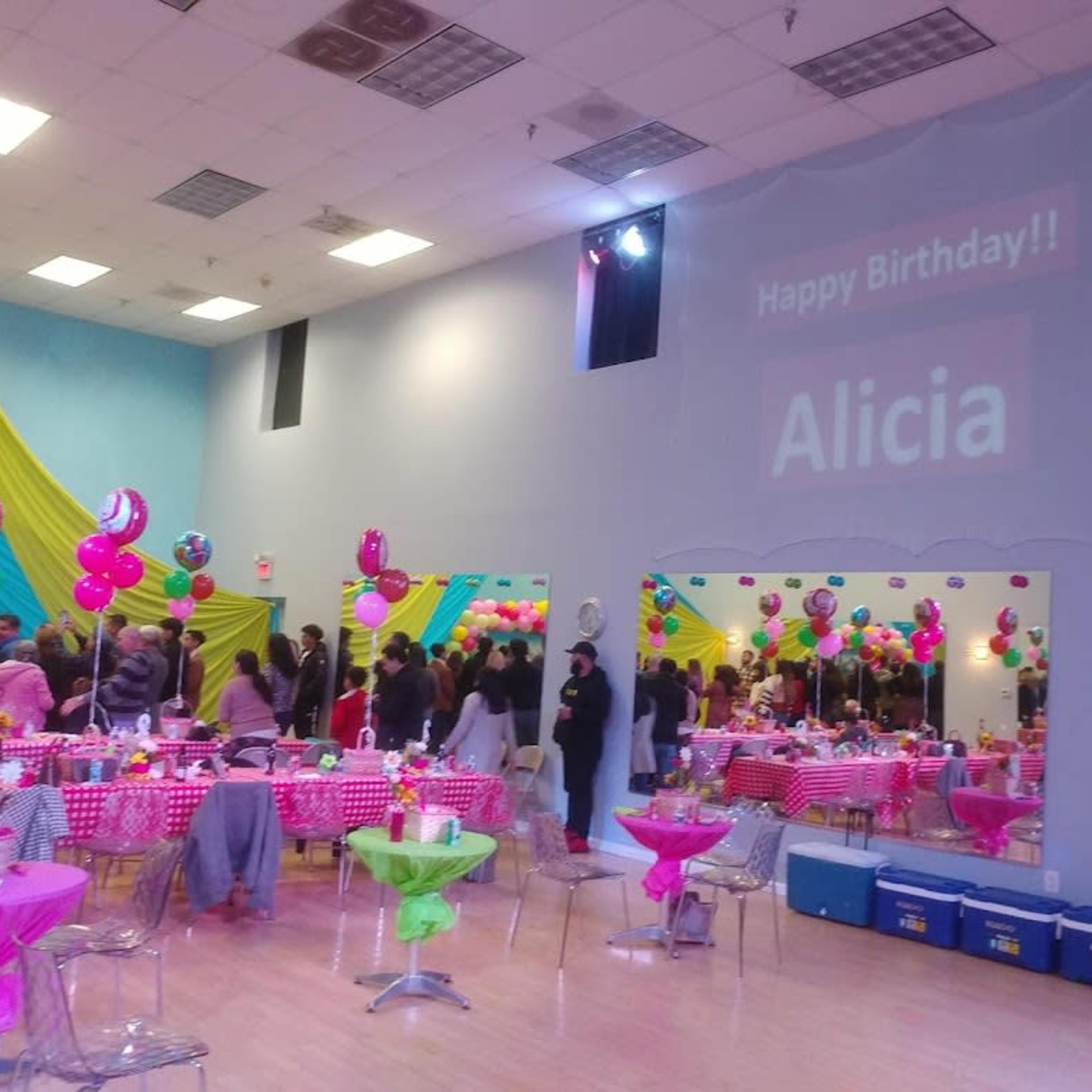 A large birthday party is set up in a colorful room with tables decorated in pink and green, and a wall display projecting "Happy Birthday!! Alicia."
