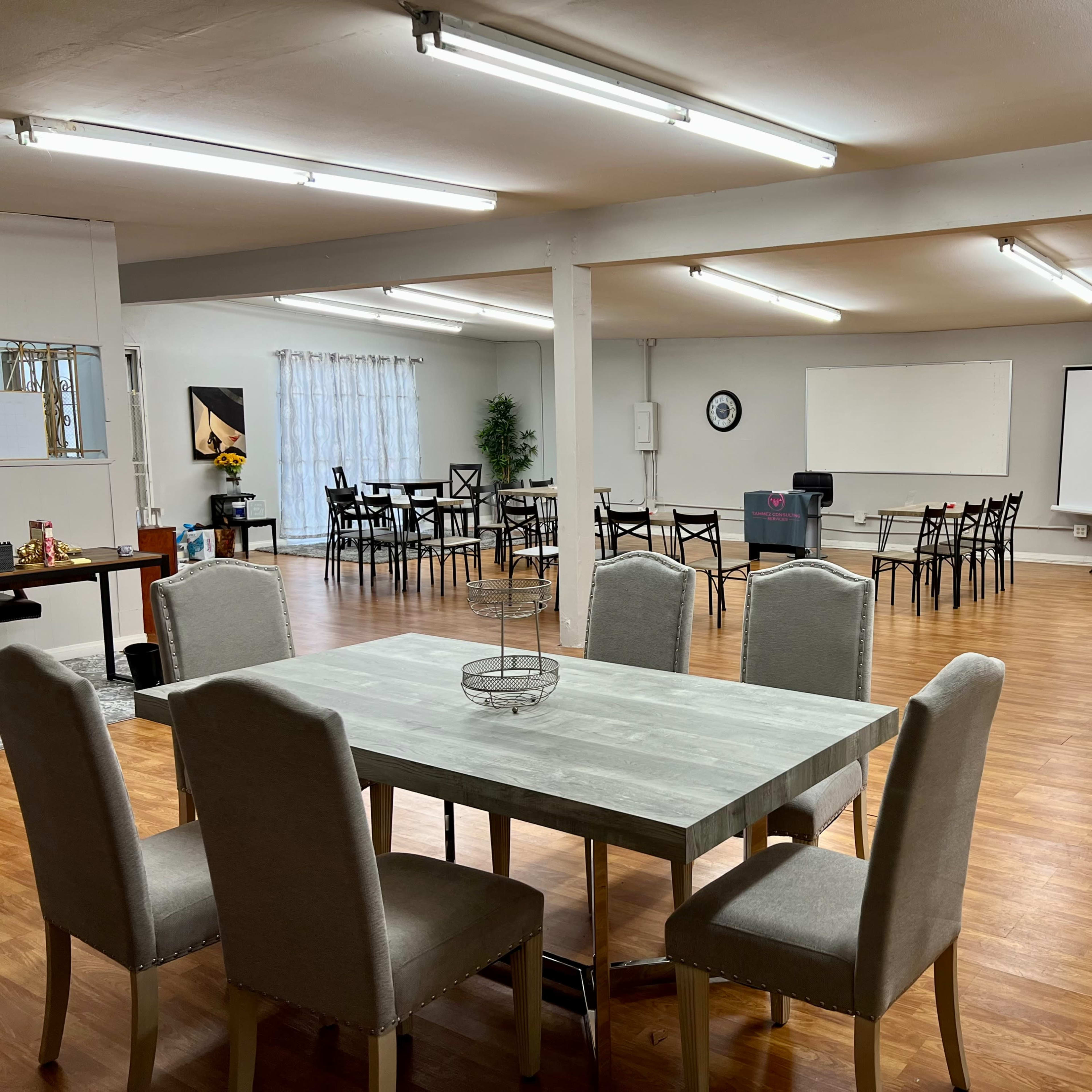 A spacious room with a dining table and chairs in the foreground, and a seating area arranged with additional chairs and a projector screen in the background.