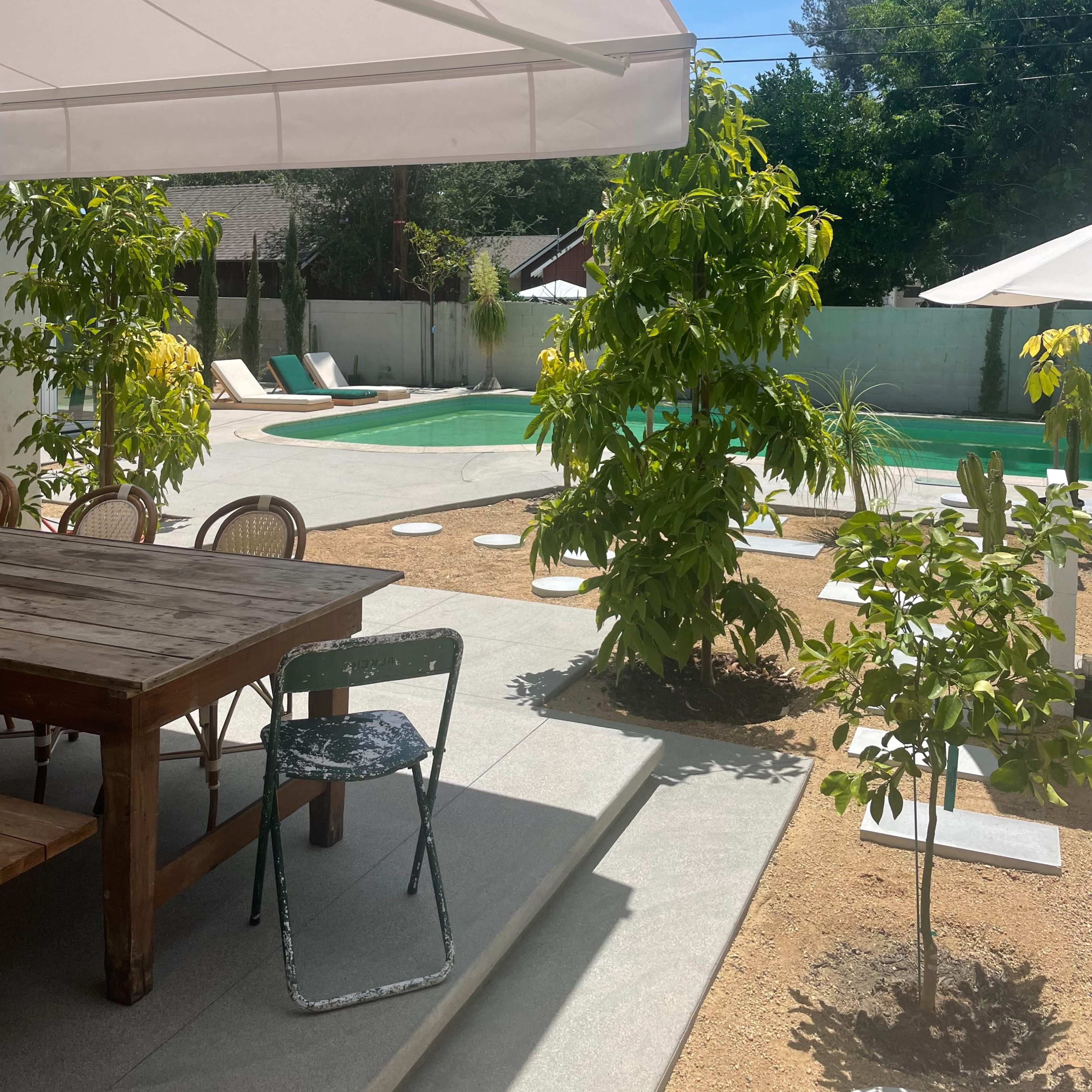 A patio area features a wooden table and chairs overlooking a swimming pool surrounded by greenery and lounge chairs.