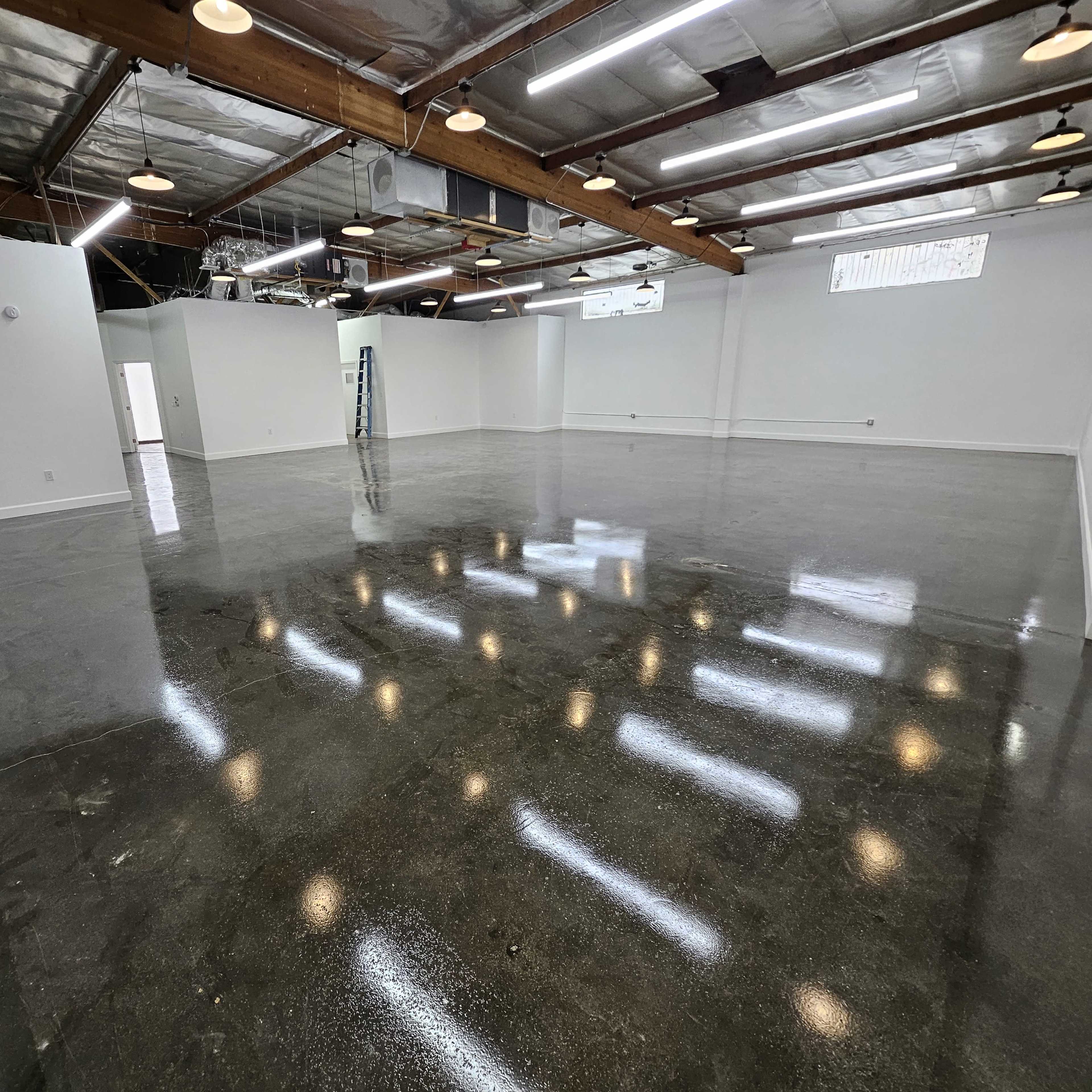 A large, empty commercial space with polished concrete floors and white walls, featuring overhead lights and a ladder in the background.