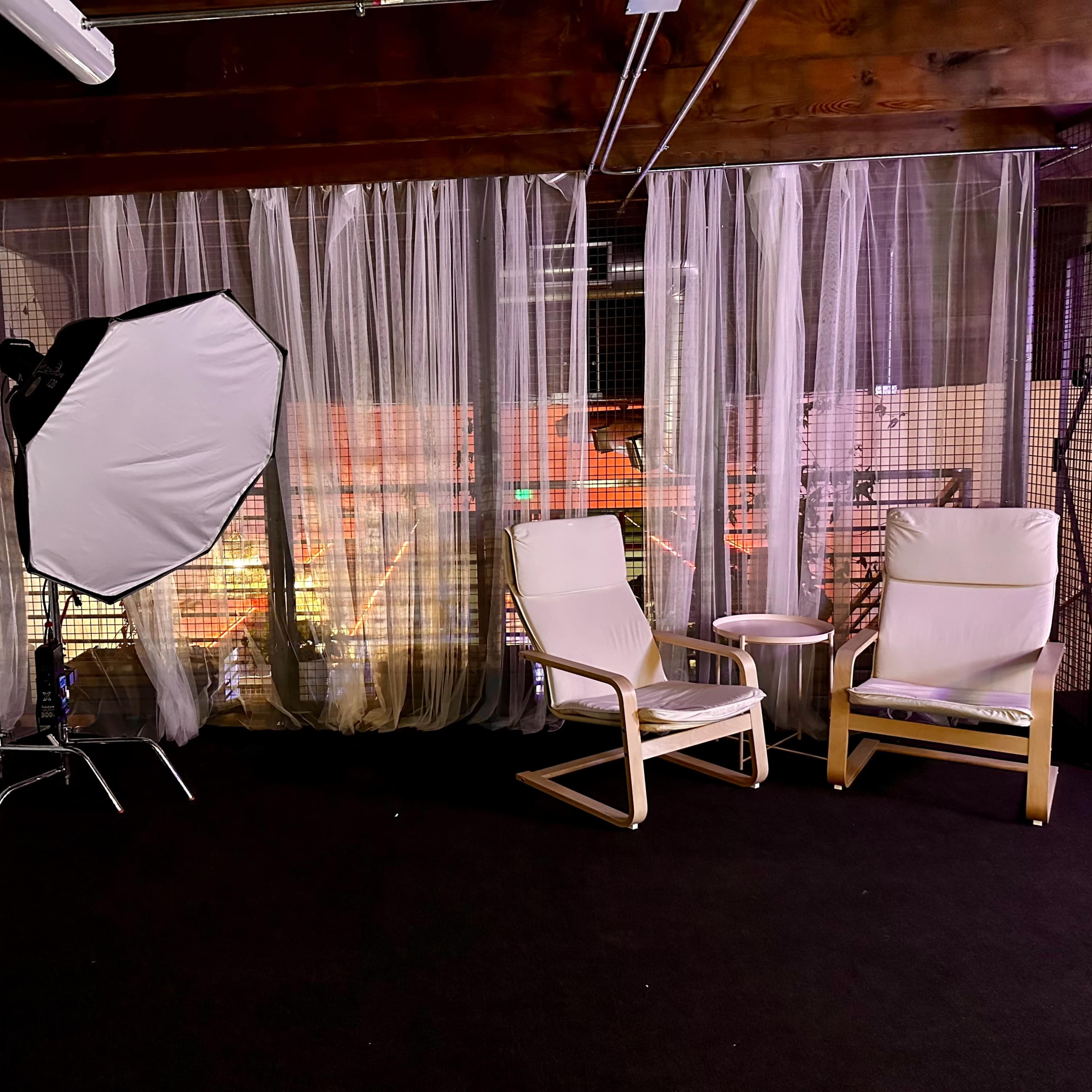 A photography studio with a softbox light, two chairs, and sheer curtains in a dimly lit space.