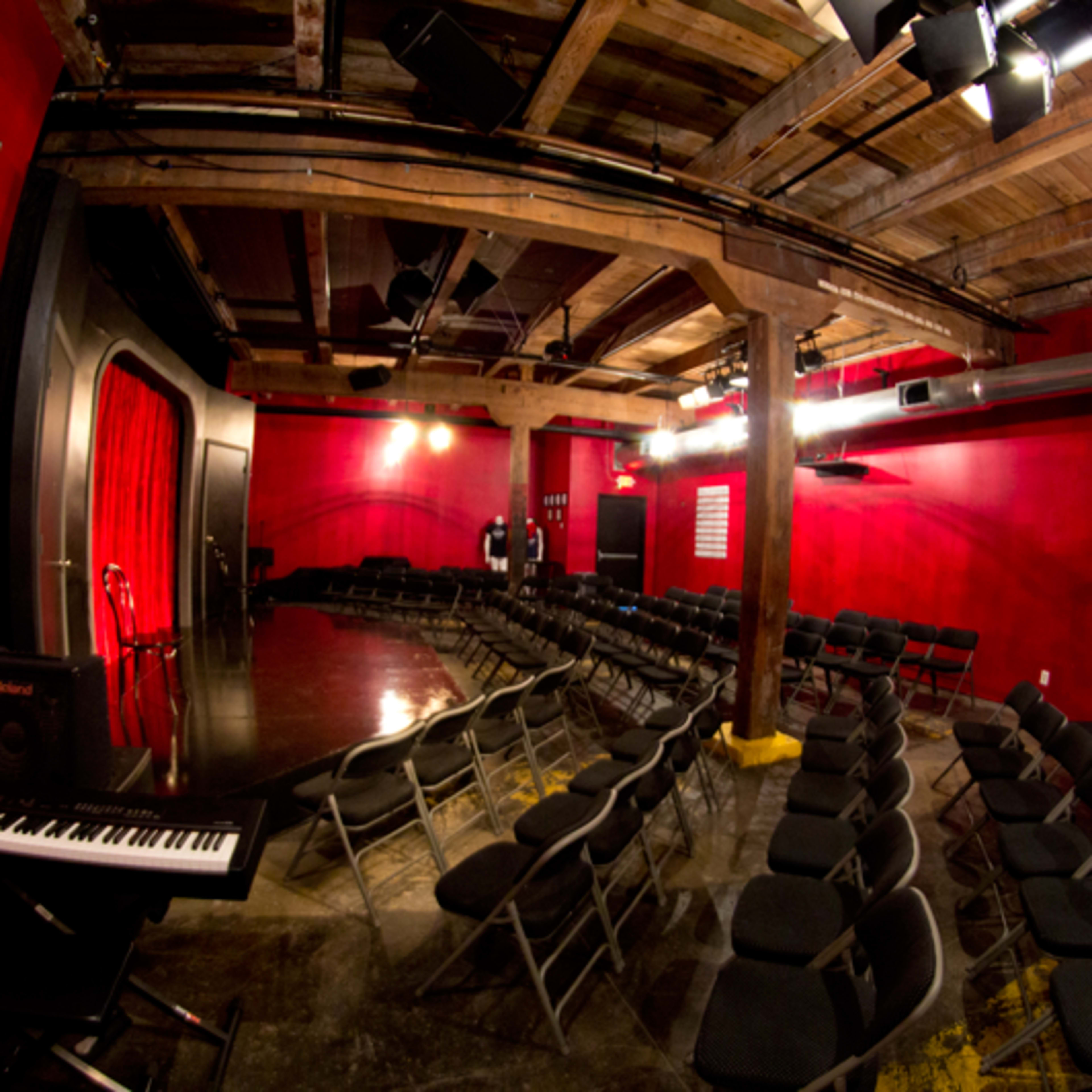 An empty comedy club with rows of black chairs facing a small stage adorned with a red curtain and a piano on the side.
