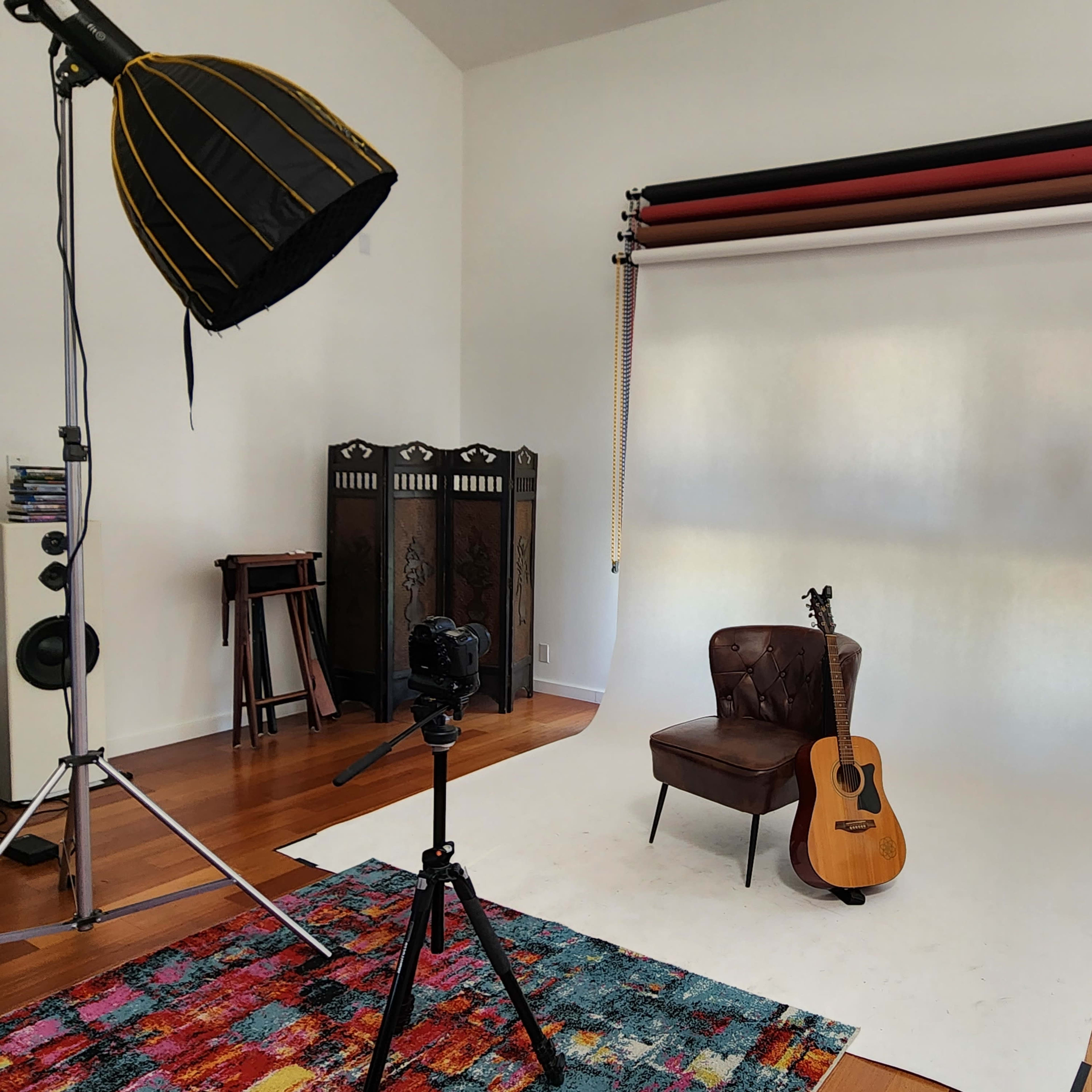A photography studio setup with a brown leather chair, an acoustic guitar, a tripods-mounted camera, and a large softbox light in a well-lit room featuring hardwood floors and white walls.