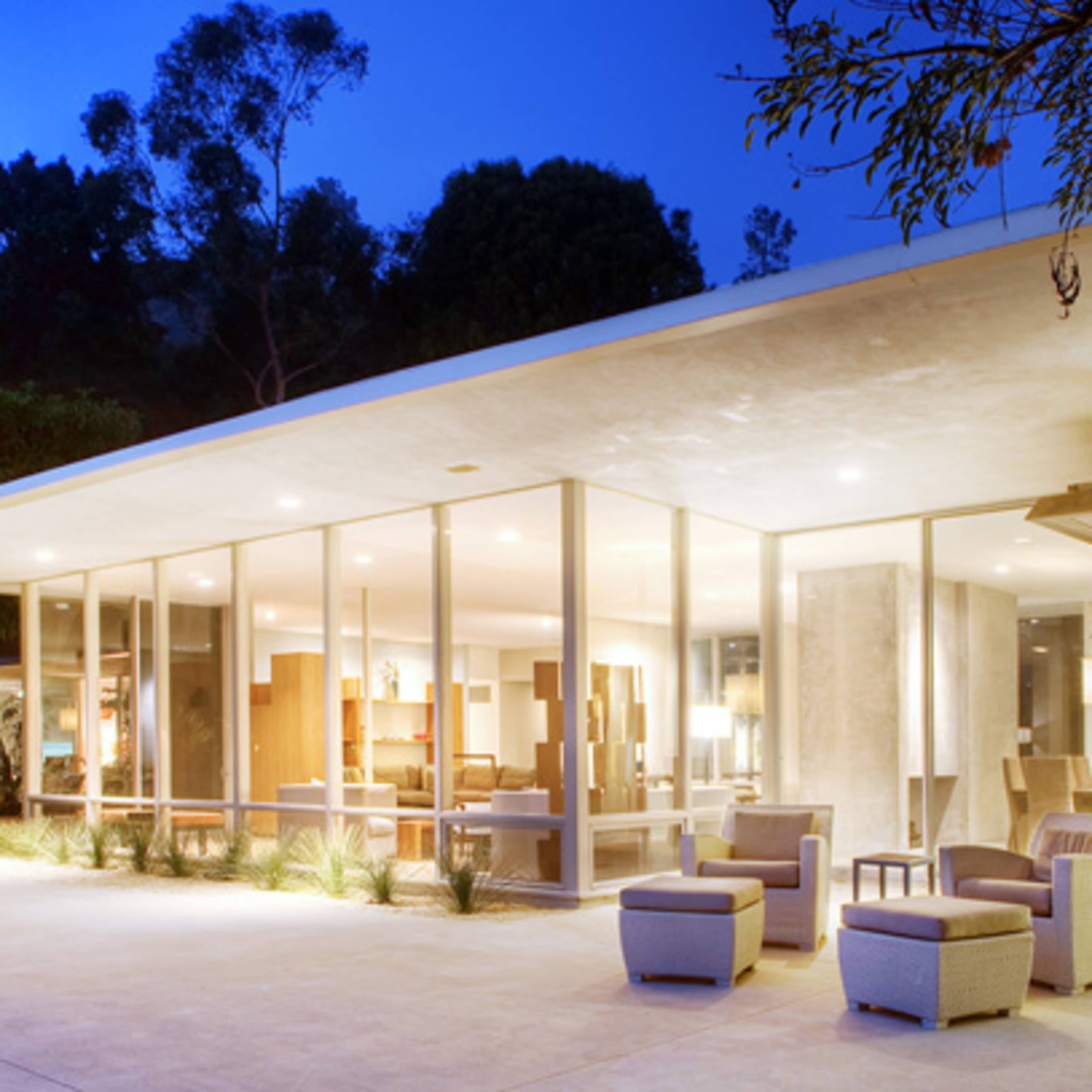 A modern house with large glass walls and a covered patio overlooks a pool and a landscaped yard in the twilight.