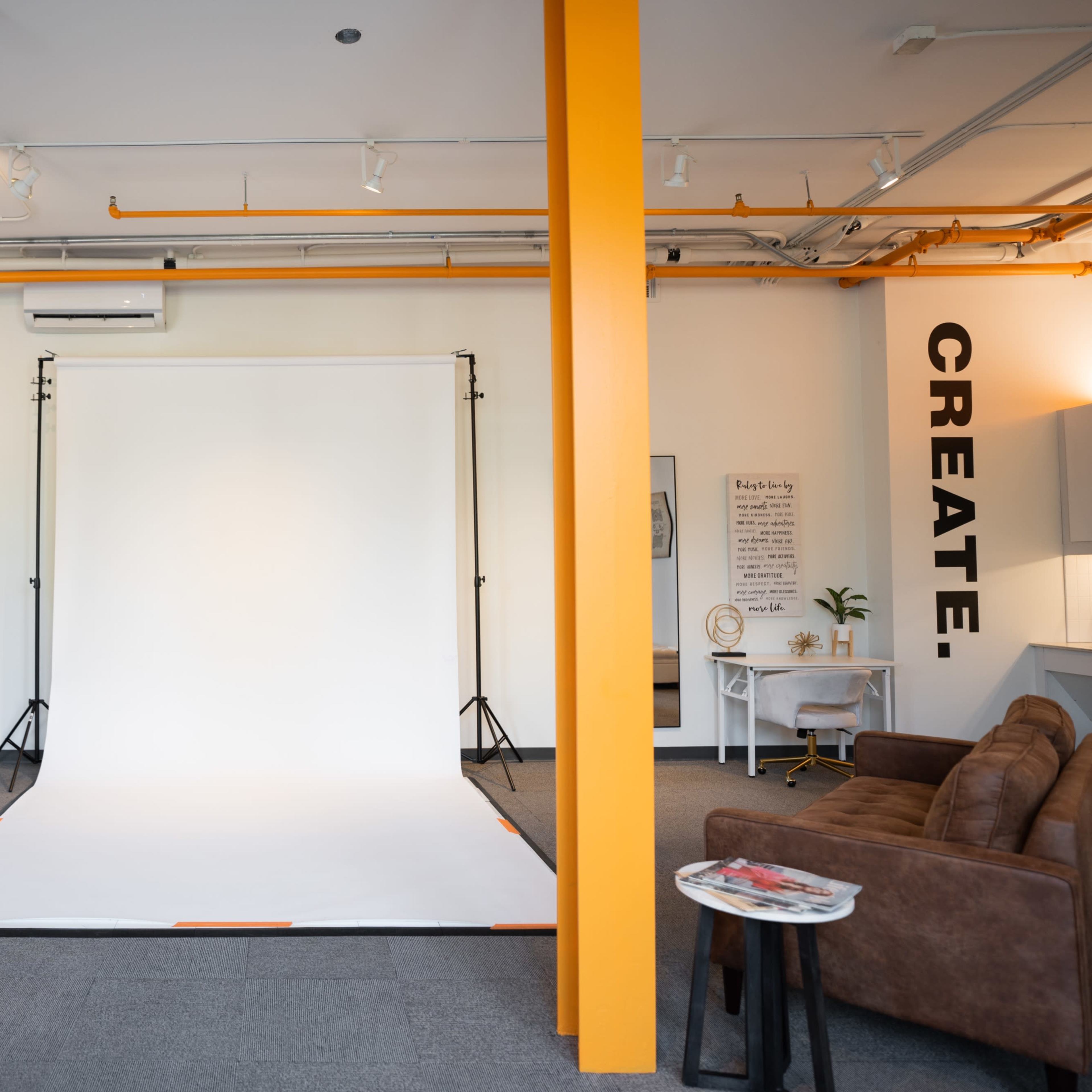A spacious studio with a white backdrop set up for photography, featuring an orange accent wall, a brown sofa, and artwork on the walls.