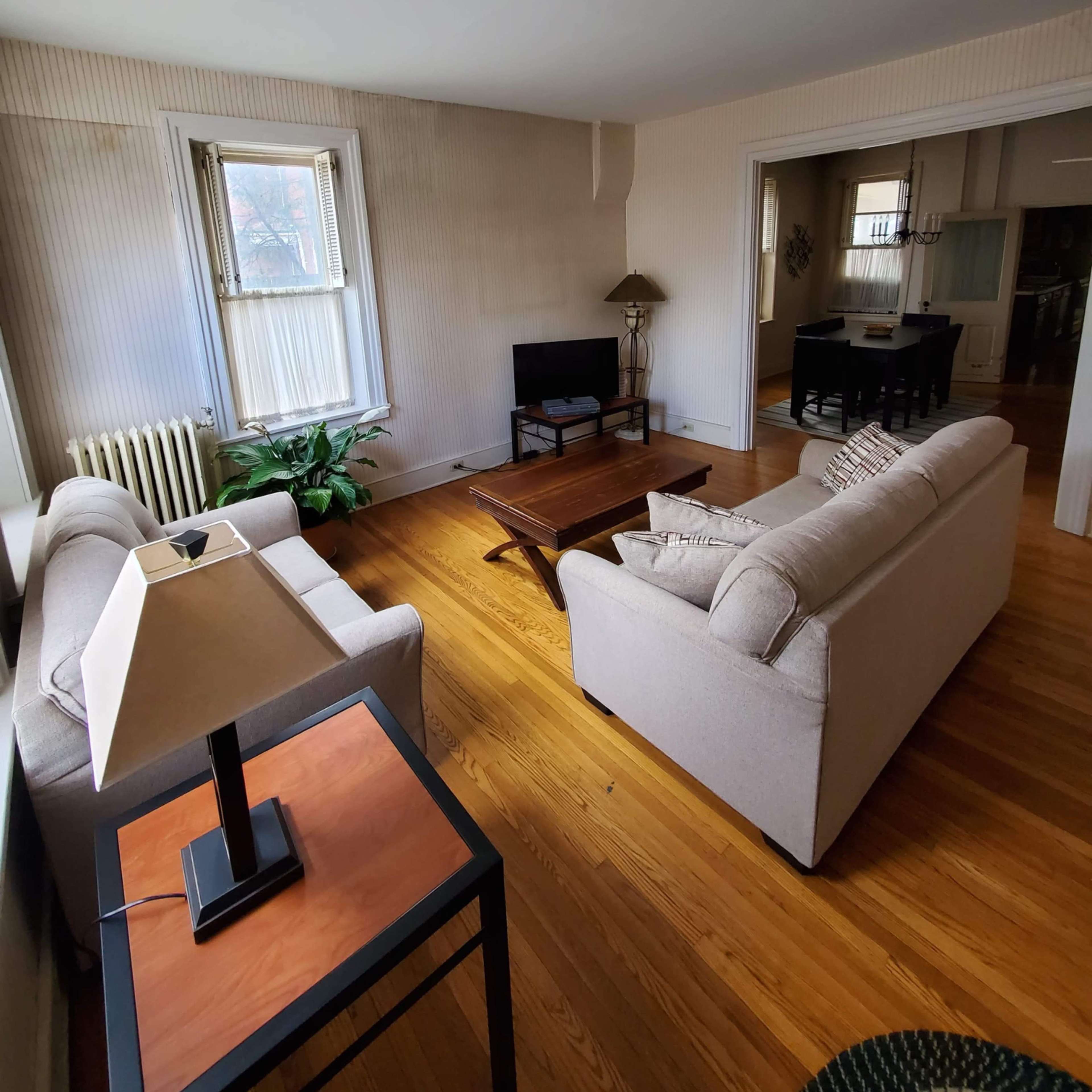 A living room with two sofas, a coffee table, a lamp, and a television, featuring hardwood floors and large windows.