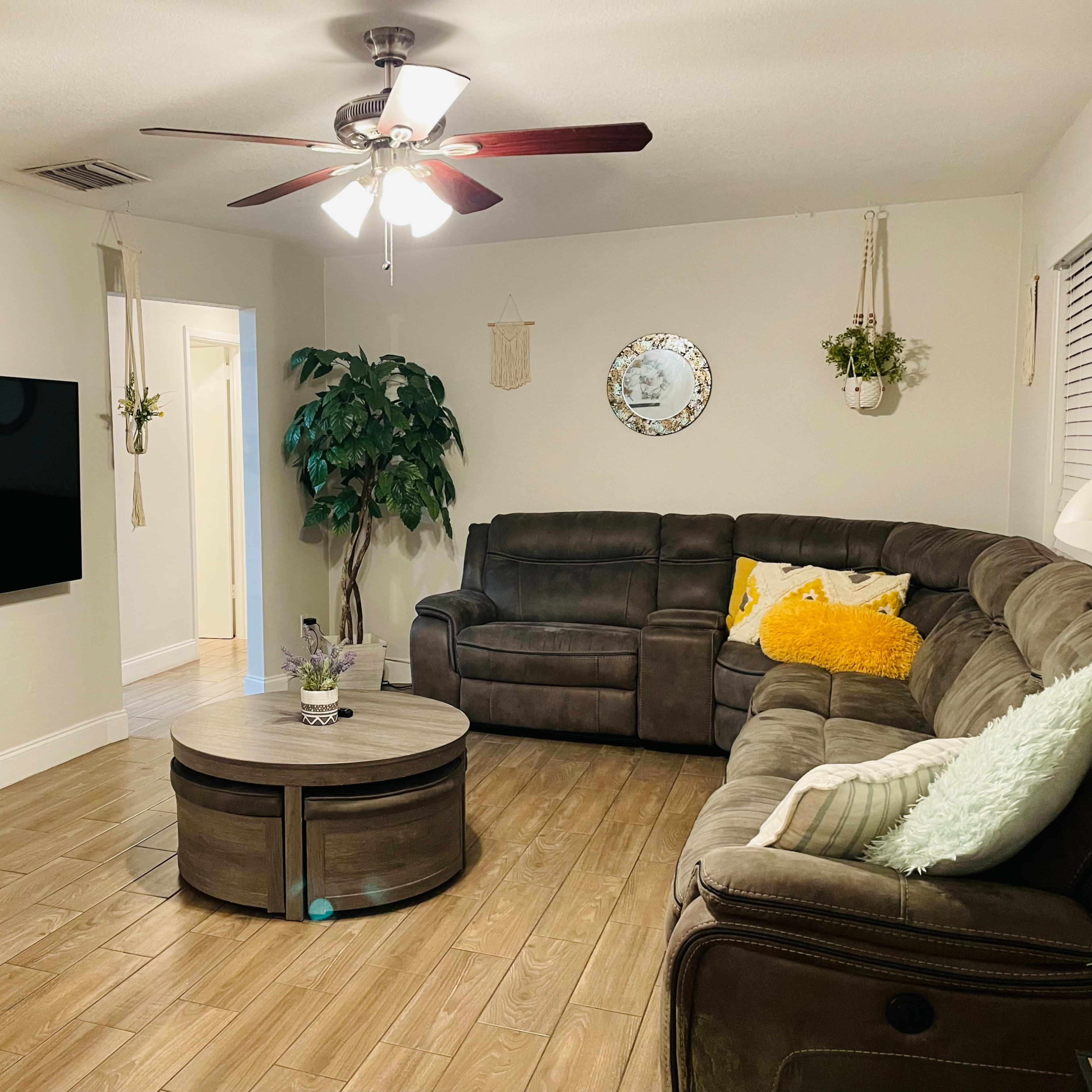 A living room with a gray sectional sofa, a round wooden coffee table, a mounted flat-screen TV, and decorative plants hanging from the walls.