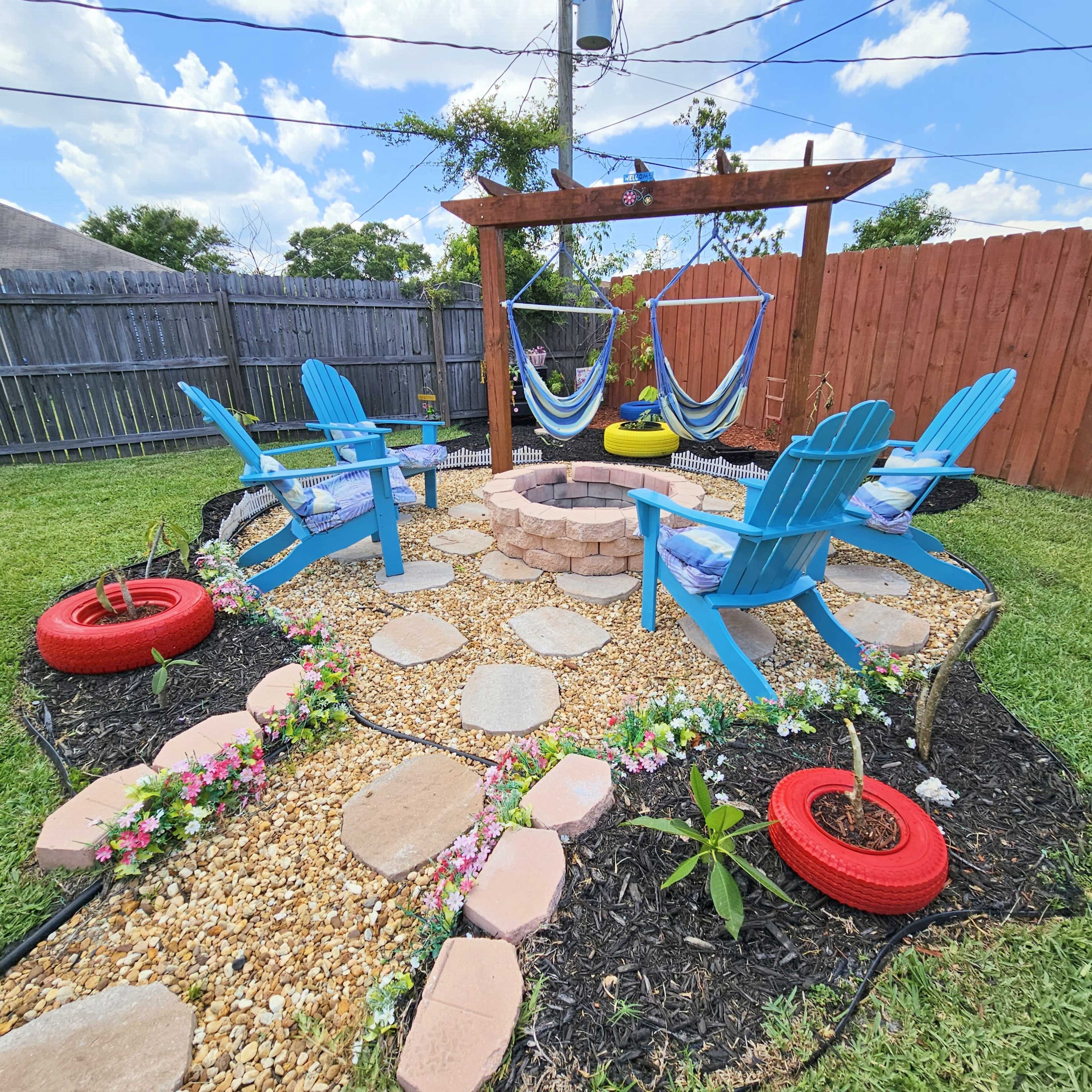 A landscaped backyard area featuring blue Adirondack chairs, a stone fire pit, and hanging hammocks, surrounded by a pathway of stones and flowers.