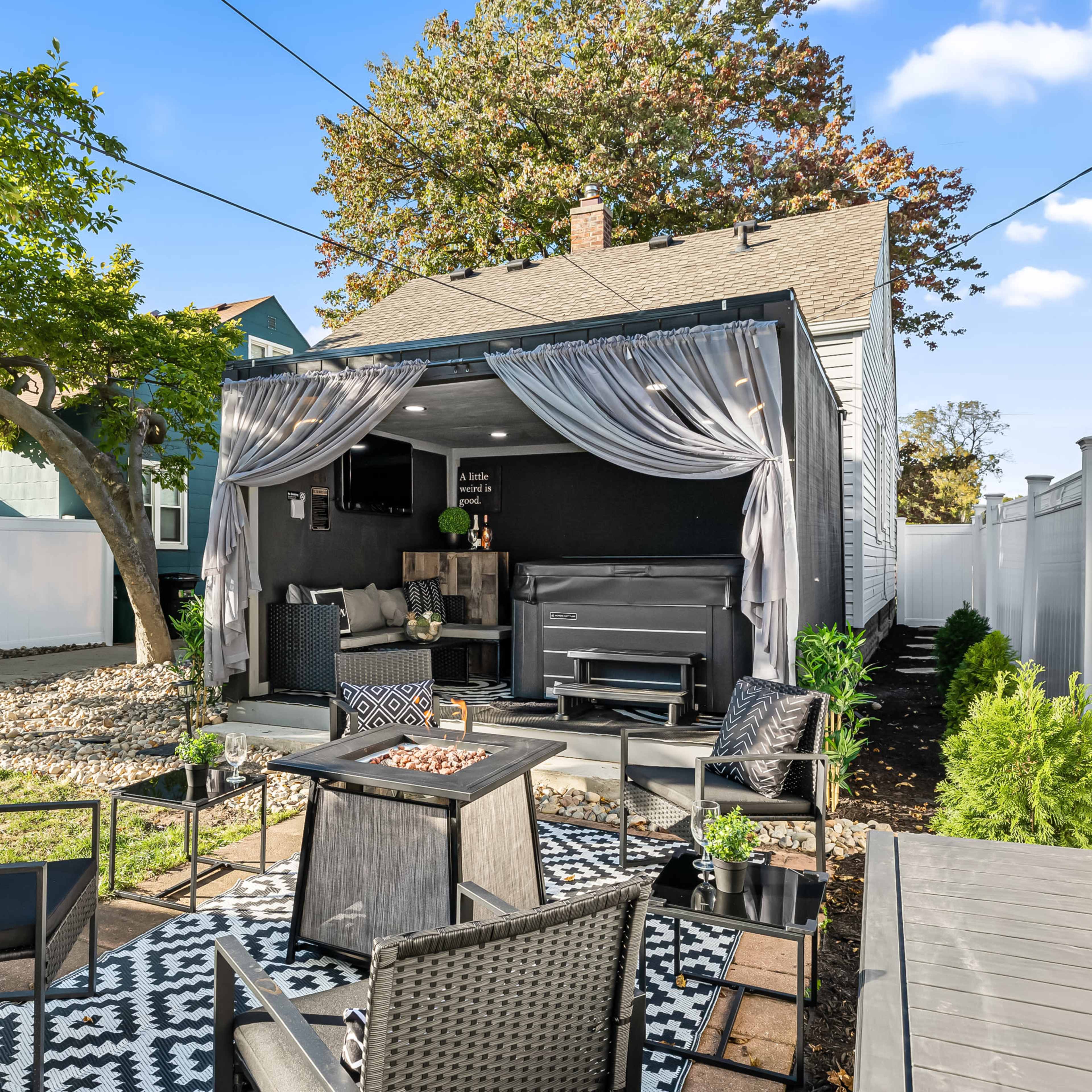 A cozy backyard patio features a covered seating area with a fire pit, surrounded by potted plants and decorative seating.