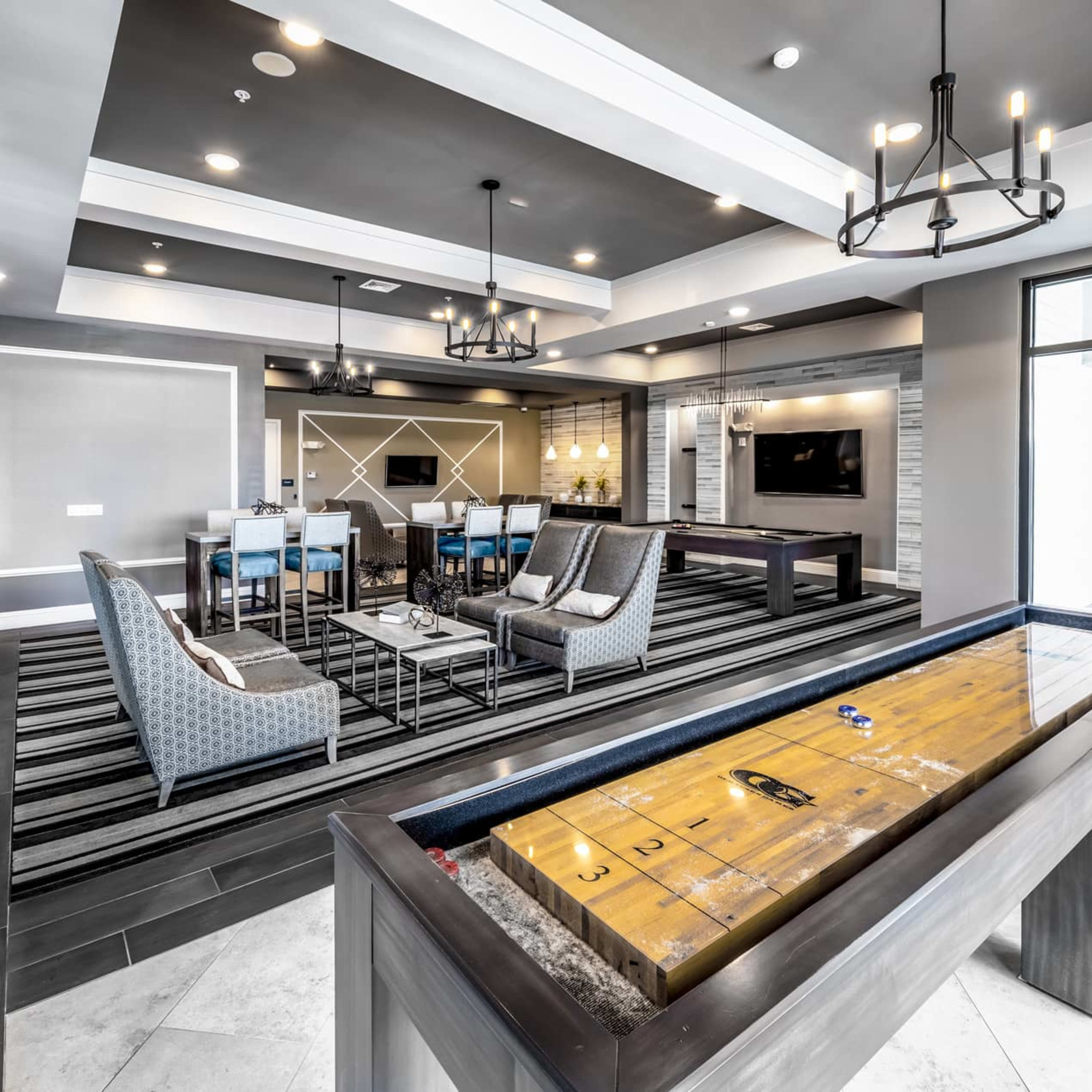 A modern lounge area with a shuffleboard table, seating arrangements, and a dining space featuring a large flat-screen TV.
