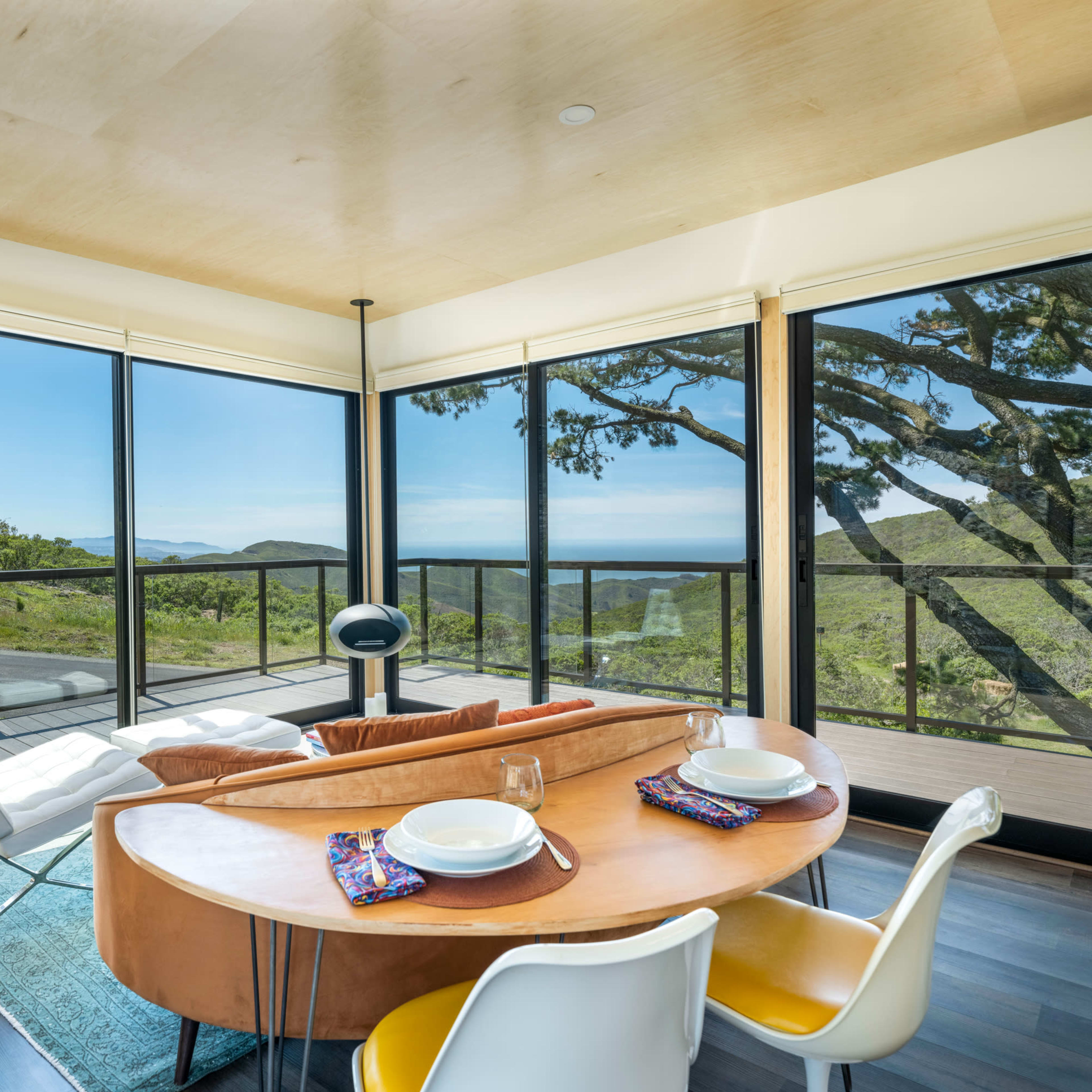 A modern dining area features a round table set for two, surrounded by large windows showcasing a view of nature and distant hills.