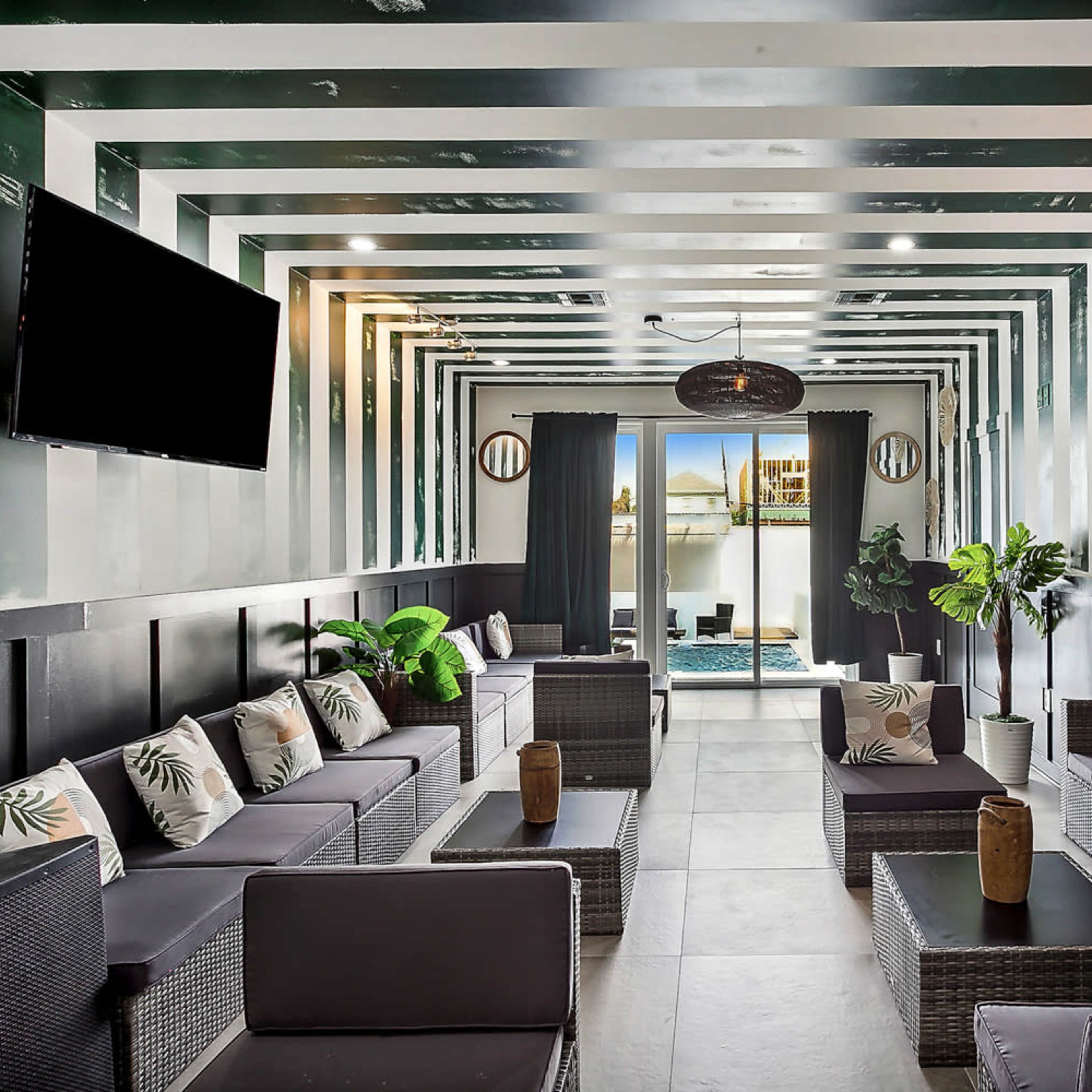 A modern lounge area with dark furniture, potted plants, and large windows overlooking a swimming pool.