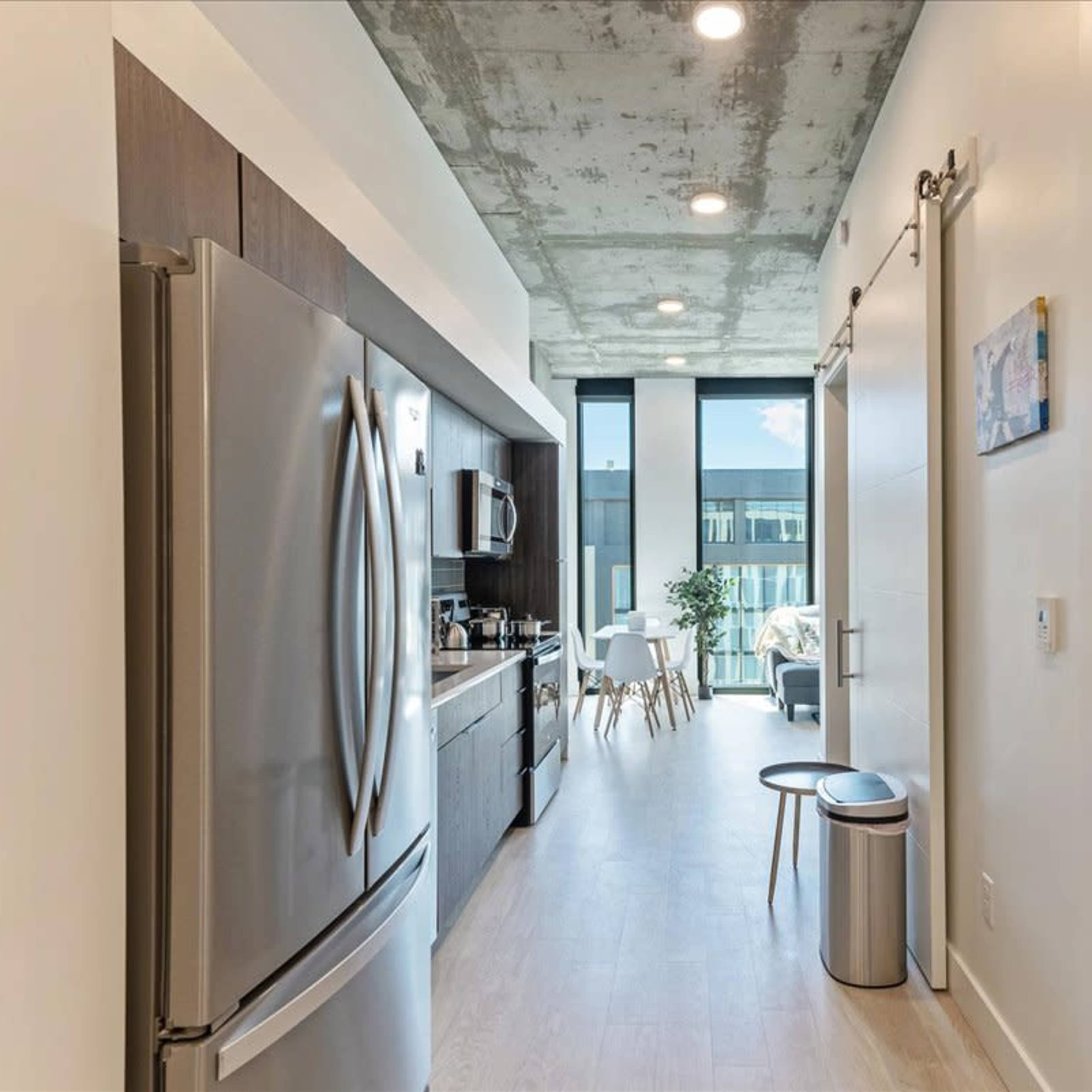 A modern kitchen and dining area are visible, featuring stainless steel appliances, a concrete ceiling, and a table with chairs near large windows.