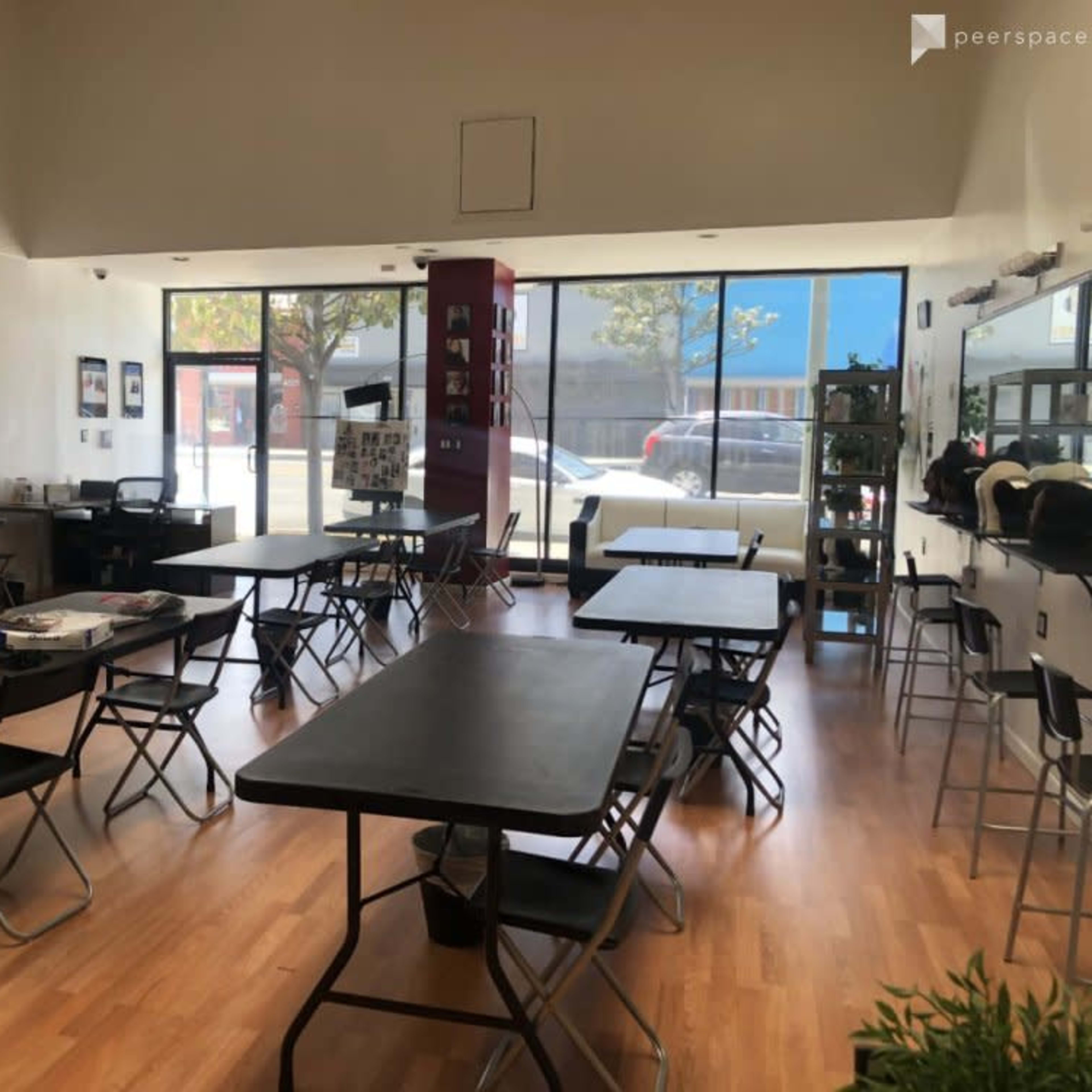 A spacious, brightly lit studio with several black tables and chairs arranged throughout, featuring large windows that allow natural light to enter.