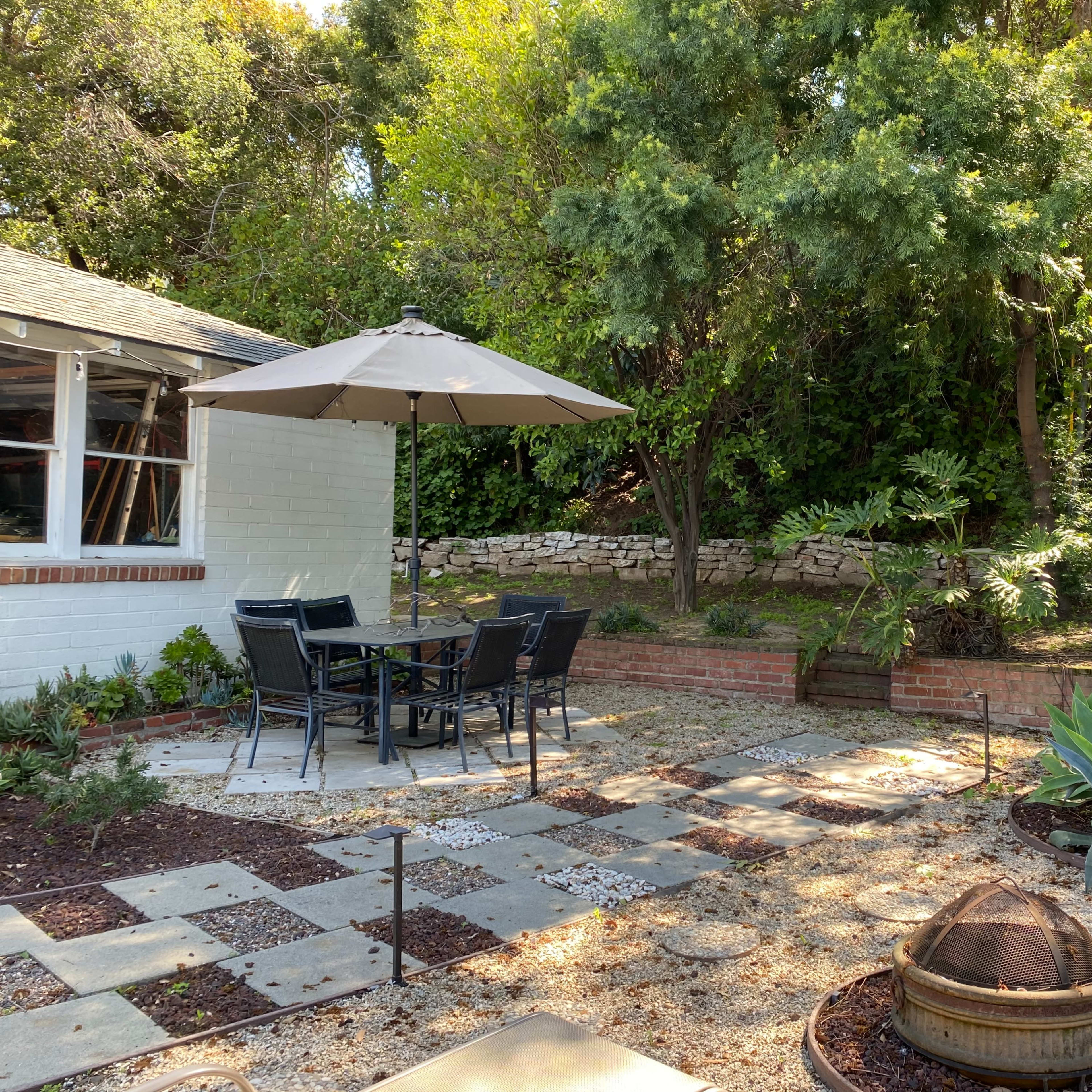 A patio area features a table with chairs beneath an umbrella, surrounded by stone pathways, planters, and various plants.