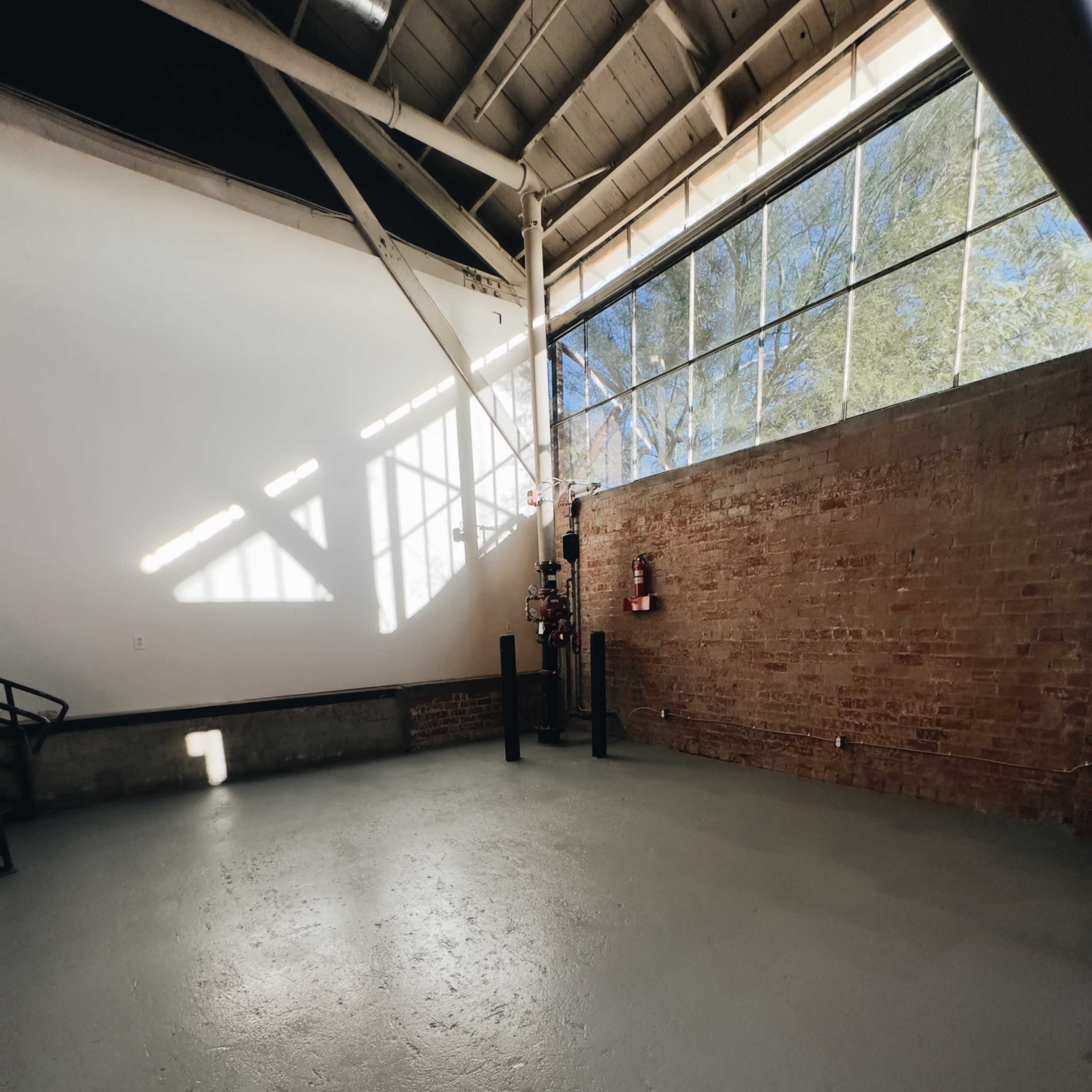 An empty room with a concrete floor, large windows allowing natural light, and exposed brick walls.