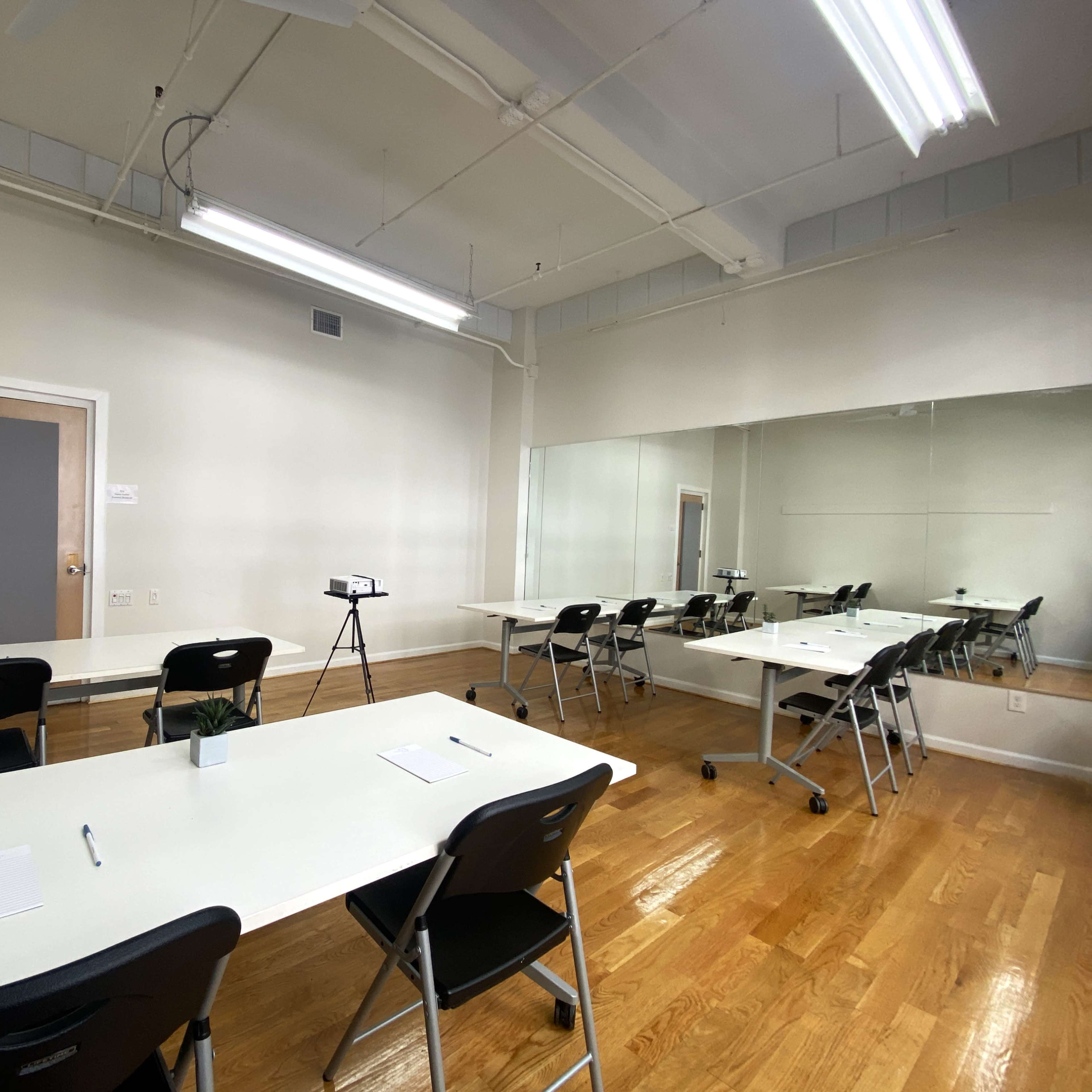 A bright, clean room with white tables and chairs arranged for a meeting, featuring a large mirror on one wall and a small plant on the table.