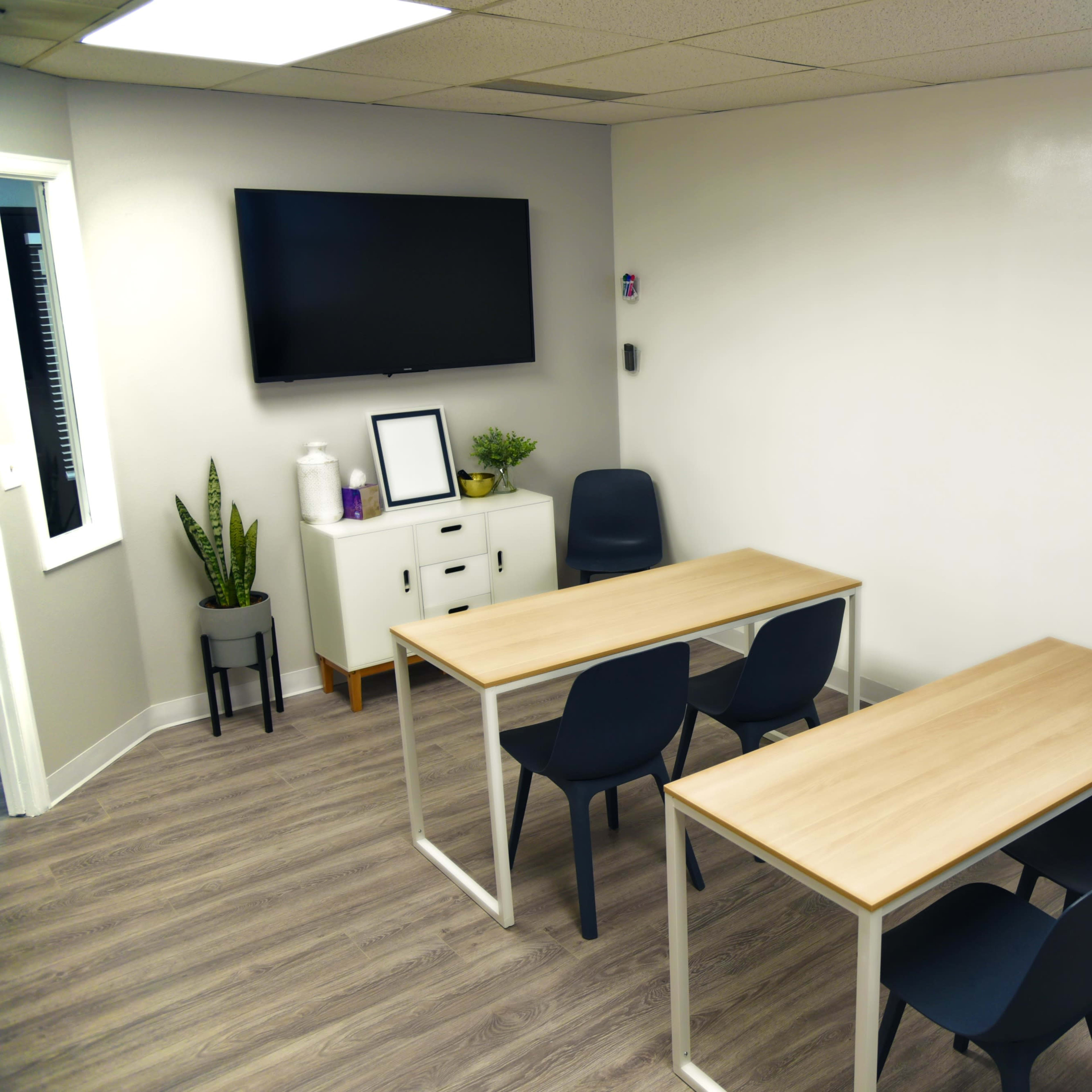 A meeting room with two wooden tables, four chairs, a wall-mounted TV, and a whiteboard.
