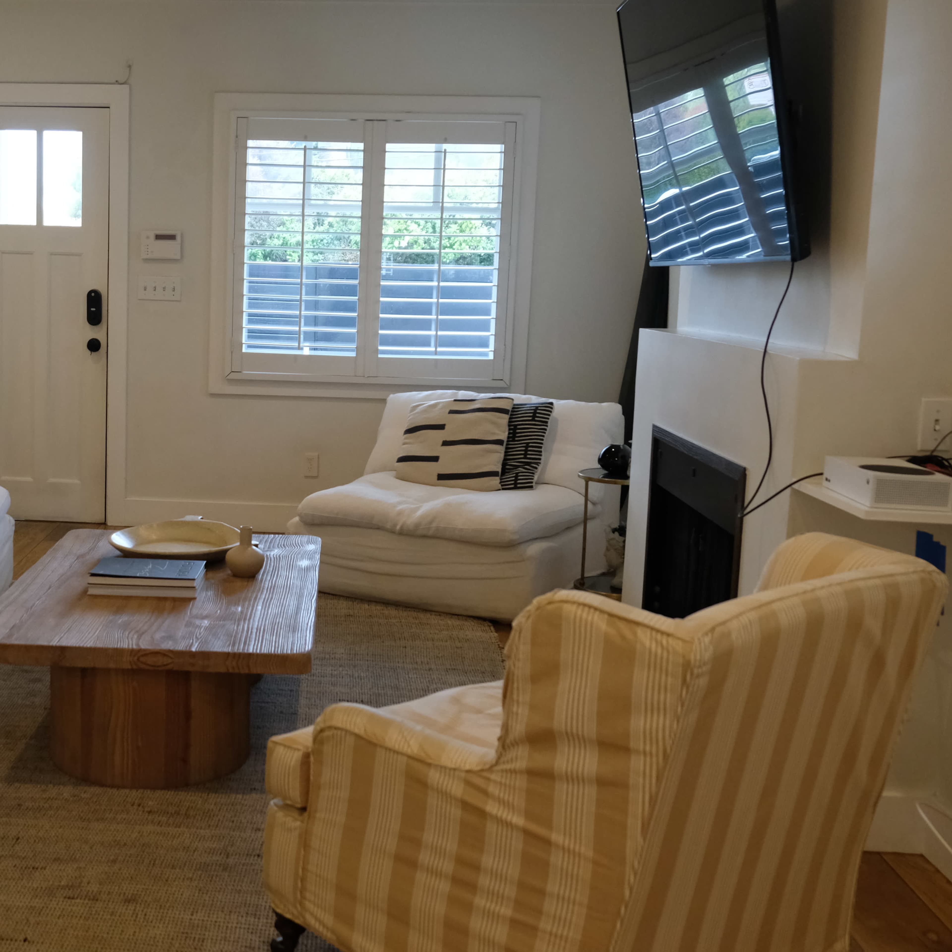 The living room features two white sofas, a wooden coffee table, and a striped armchair, with a television mounted on the wall and sunlight streaming through the window.