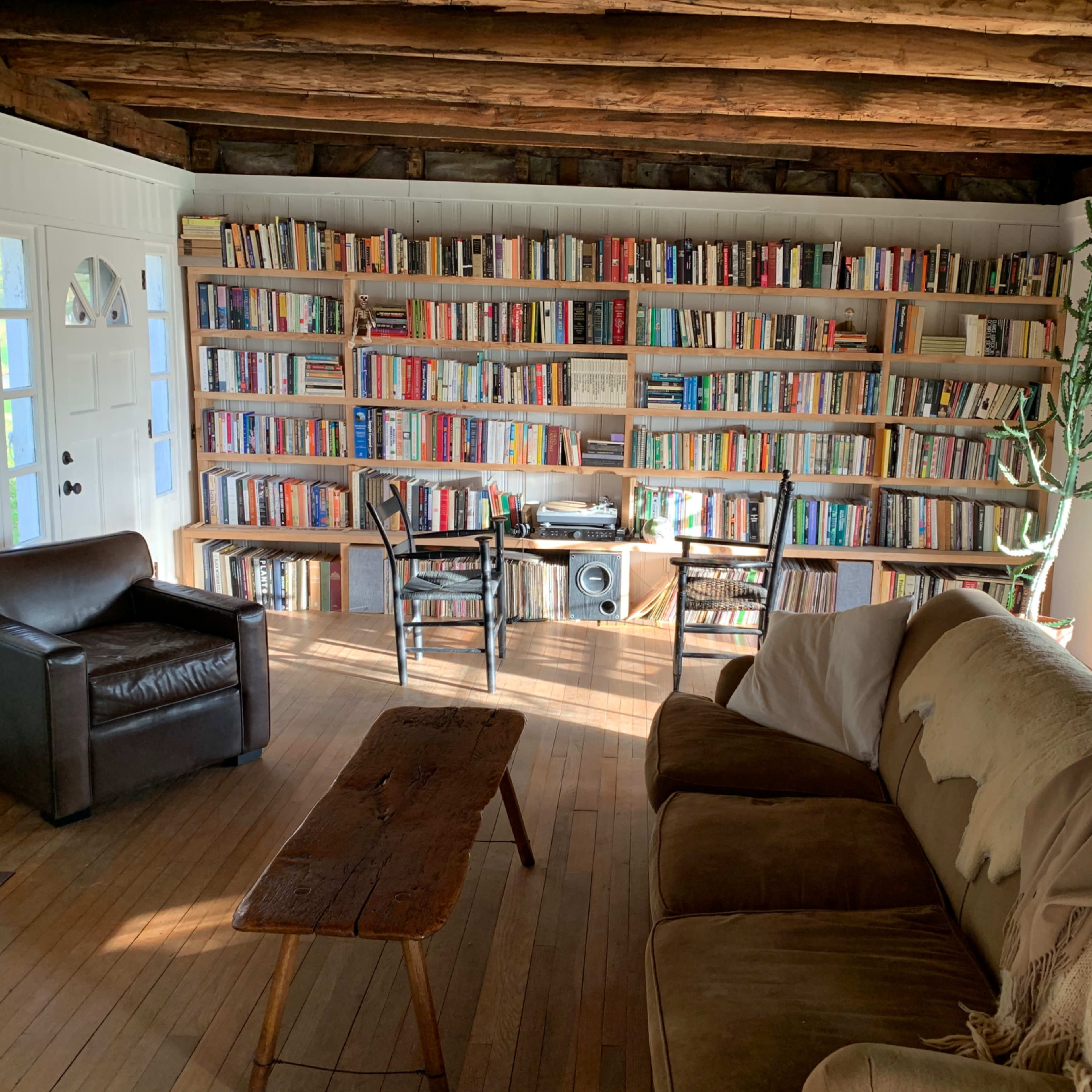 A cozy living space features a large bookshelf filled with books, a wooden coffee table, two chairs, and a sectional sofa with a throw blanket, illuminated by natural light.