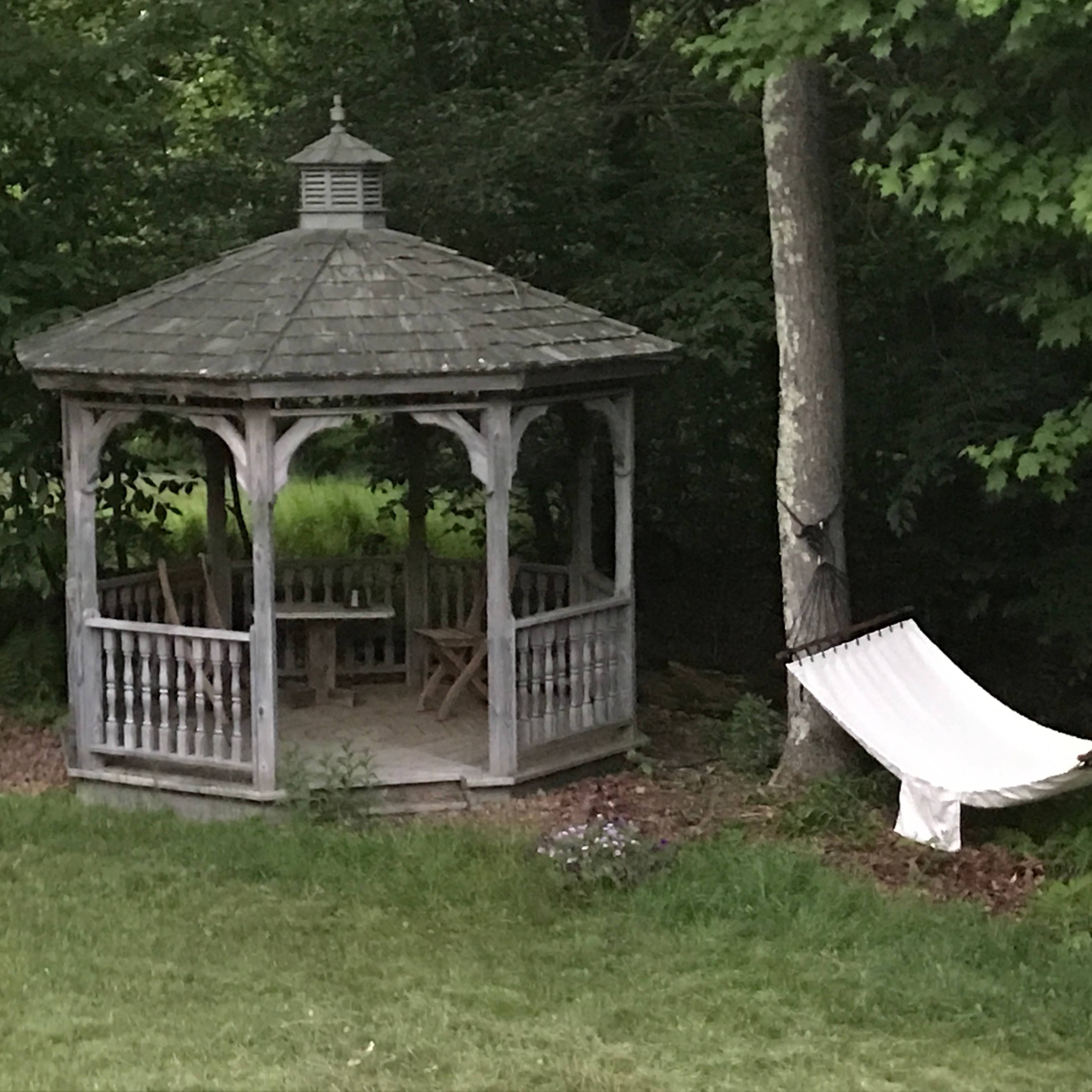 A wooden gazebo with a sitting area is located in a grassy area beside a hammock hanging between two trees.