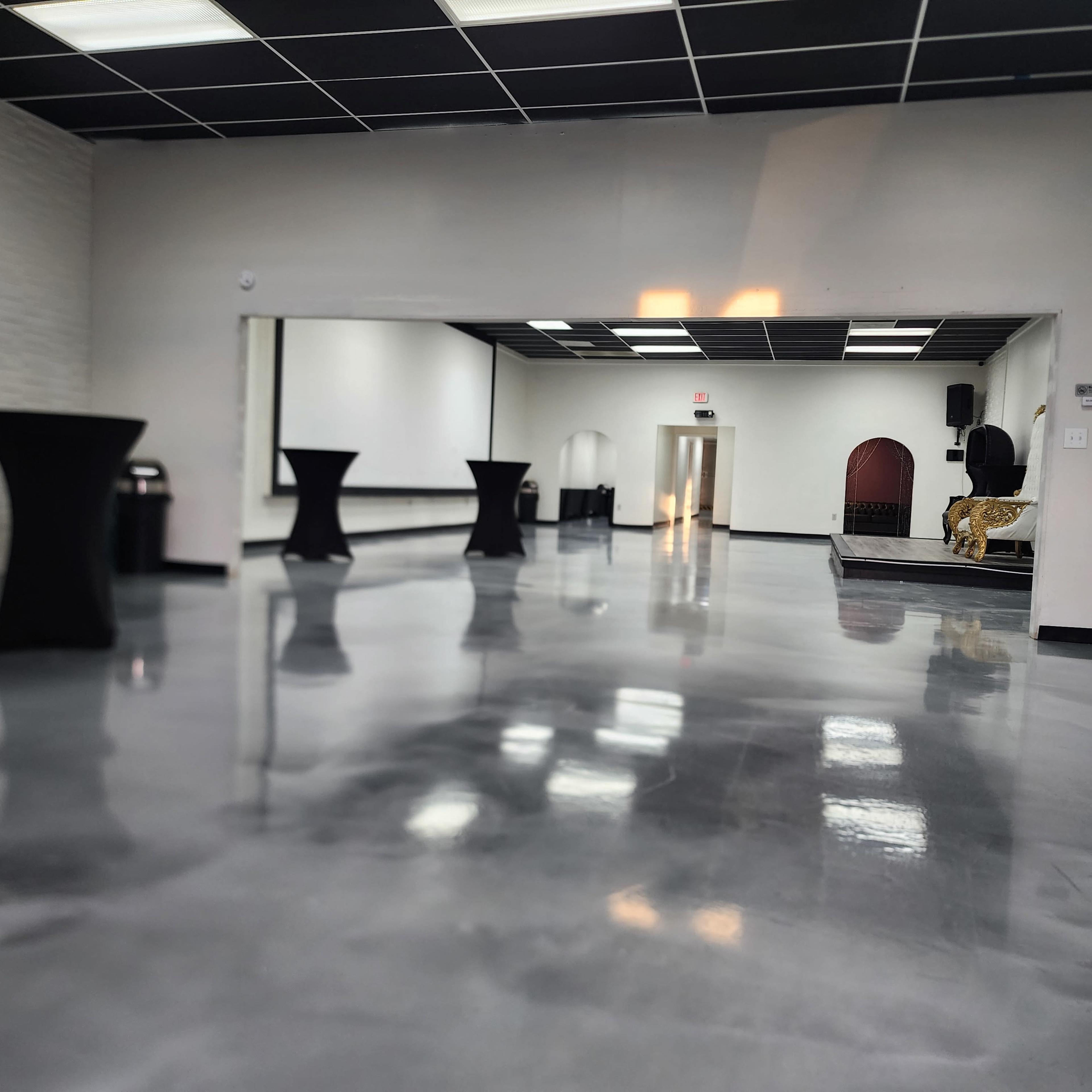 An empty modern event space with polished floors, tall black tables, and an elevated stage at the far end.
