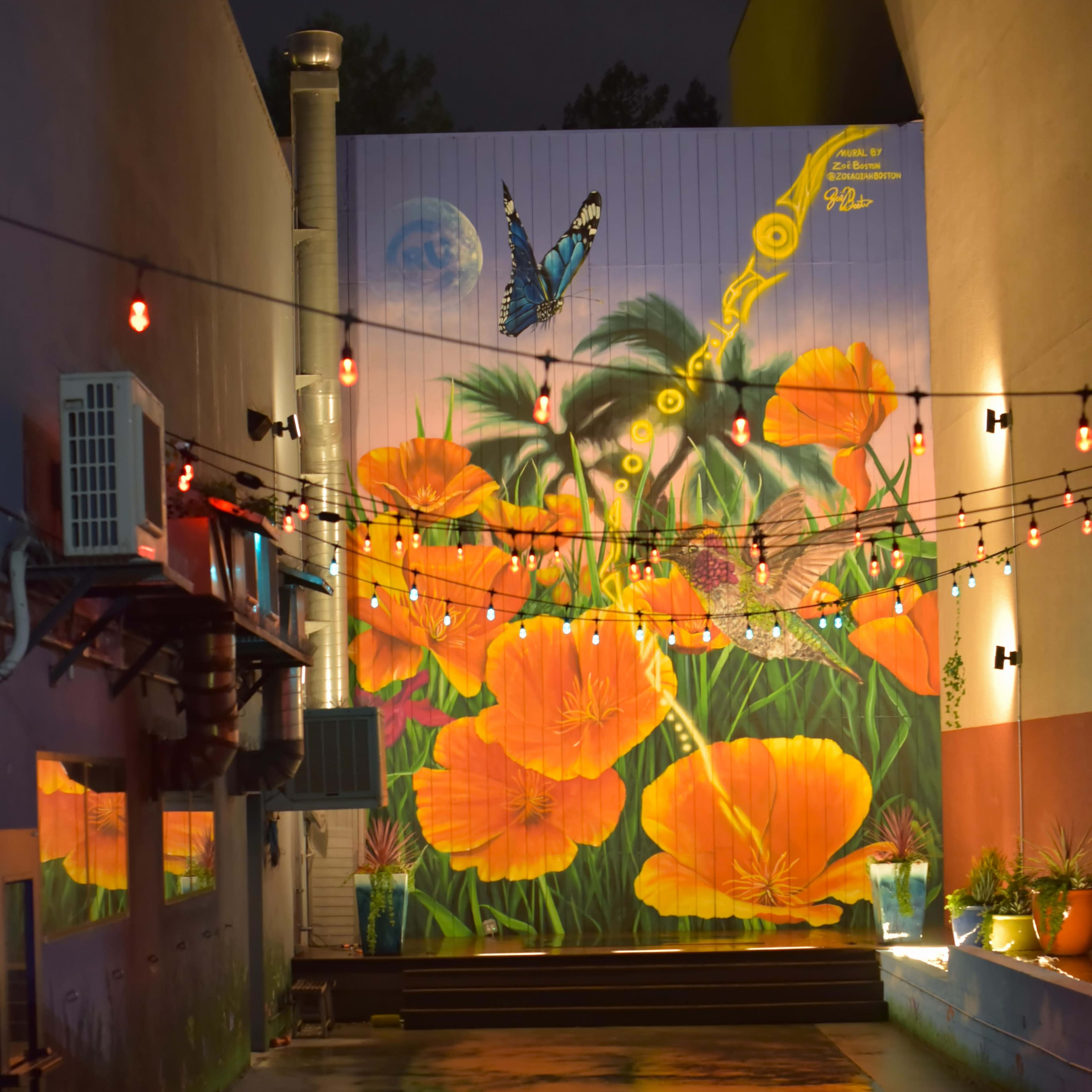 A brightly painted mural of orange flowers and a butterfly decorates a wall in a dimly lit alley, reflecting on a wet surface below.