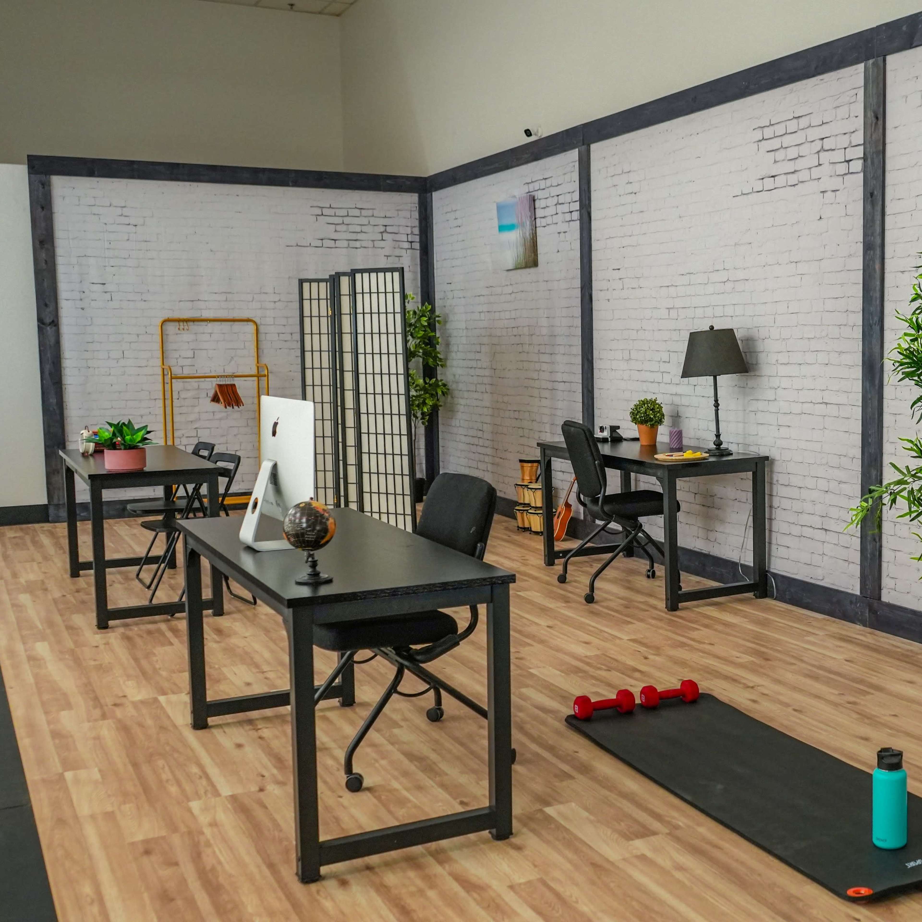 A modern, minimalist workspace featuring two desks, plants, exercise equipment, and a blank easel against a white brick wall.