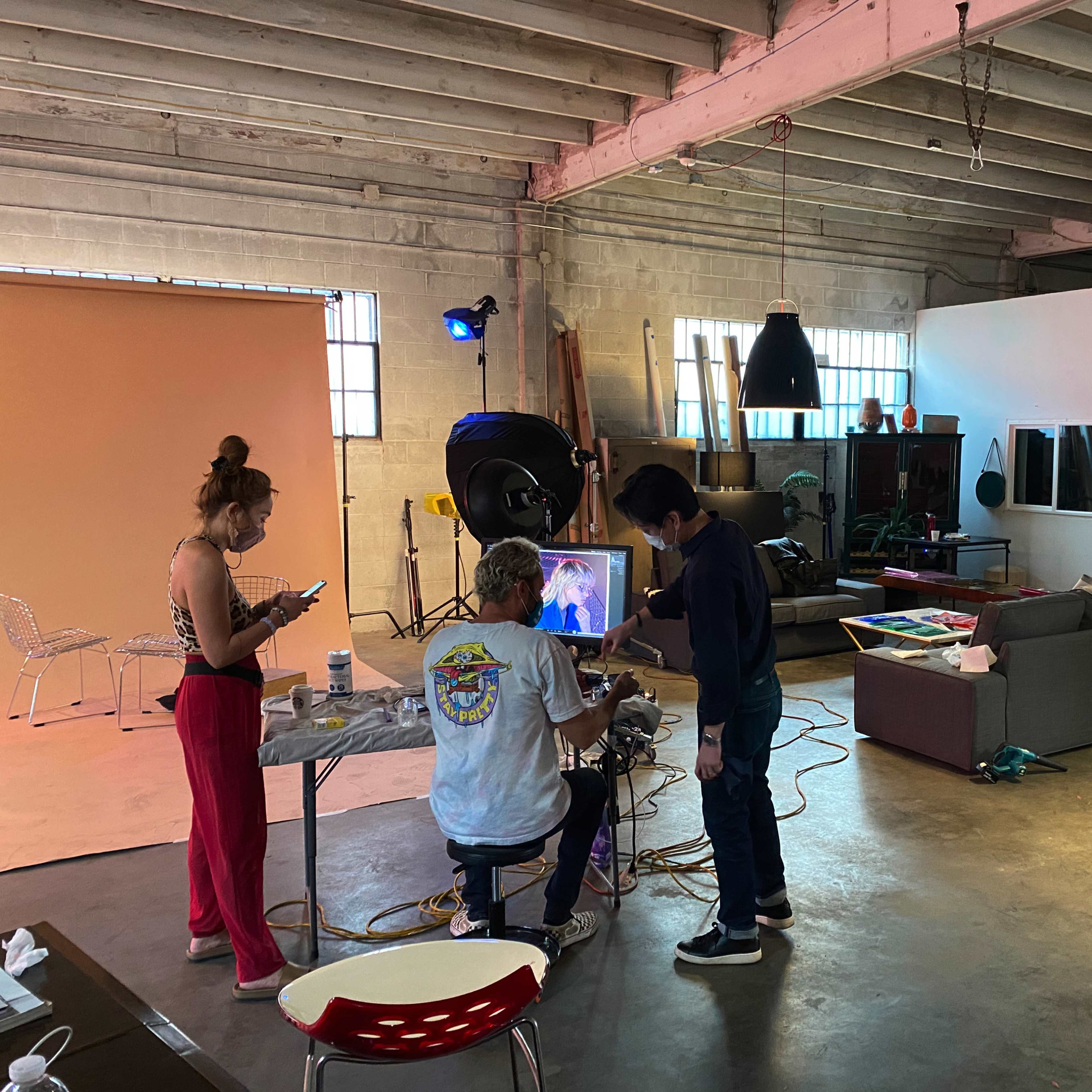 A studio space is set up for a photoshoot, featuring a model preparing in a chair, while a photographer adjusts equipment and another person uses a phone nearby.