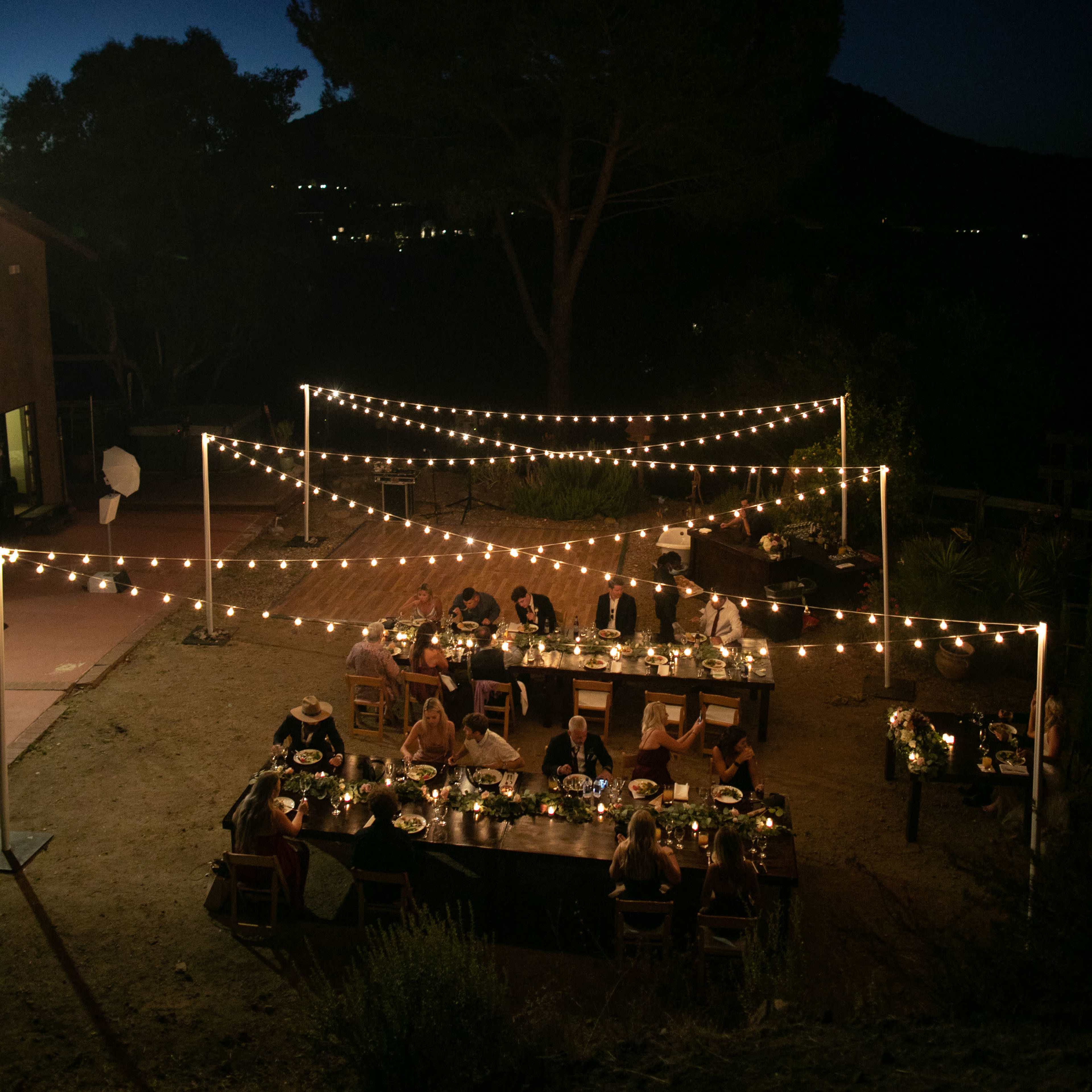 A long outdoor dining table is set for a dinner party under string lights on a clear night.