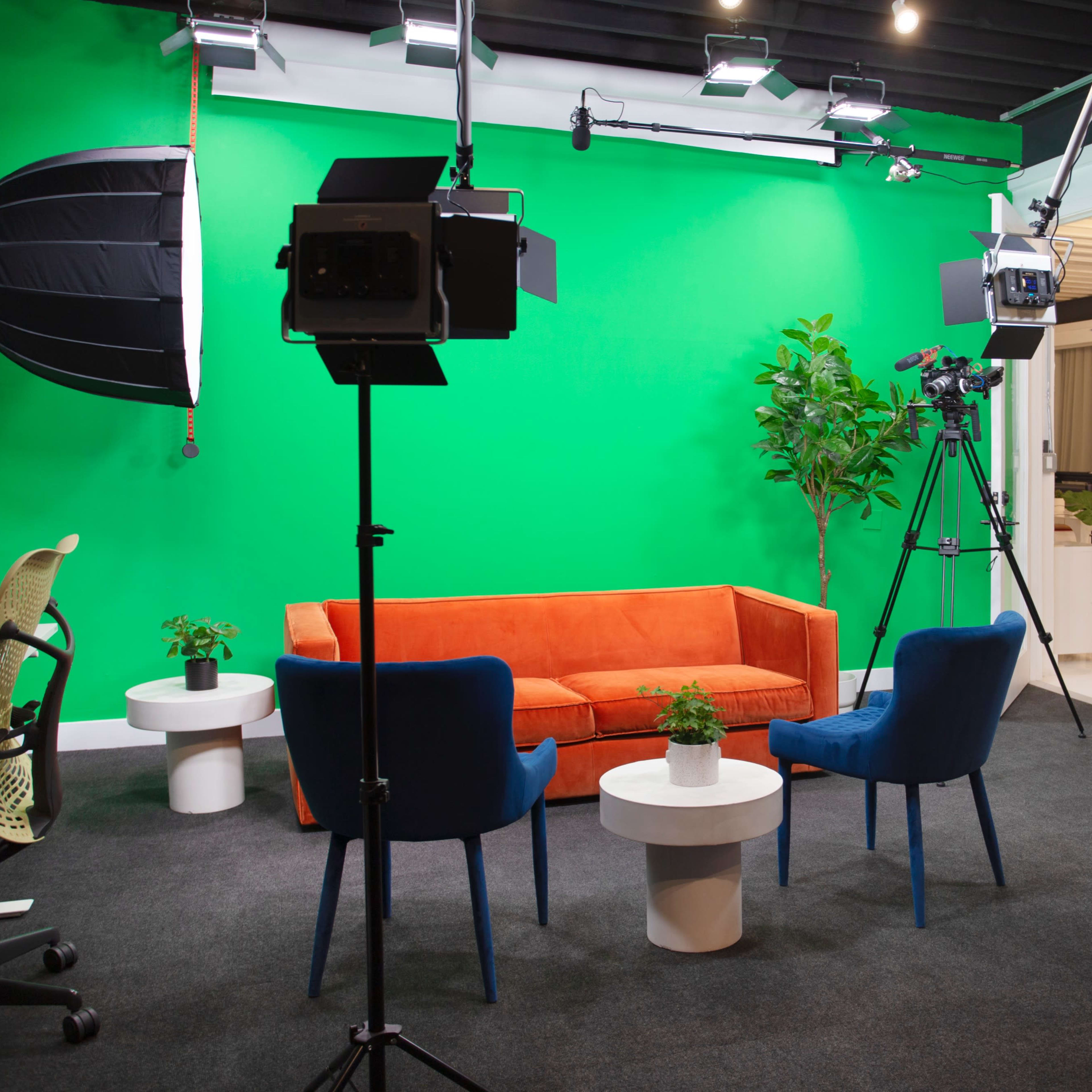 A studio setup with a green backdrop, an orange couch, two blue chairs, small round tables, and camera equipment positioned to capture the scene.