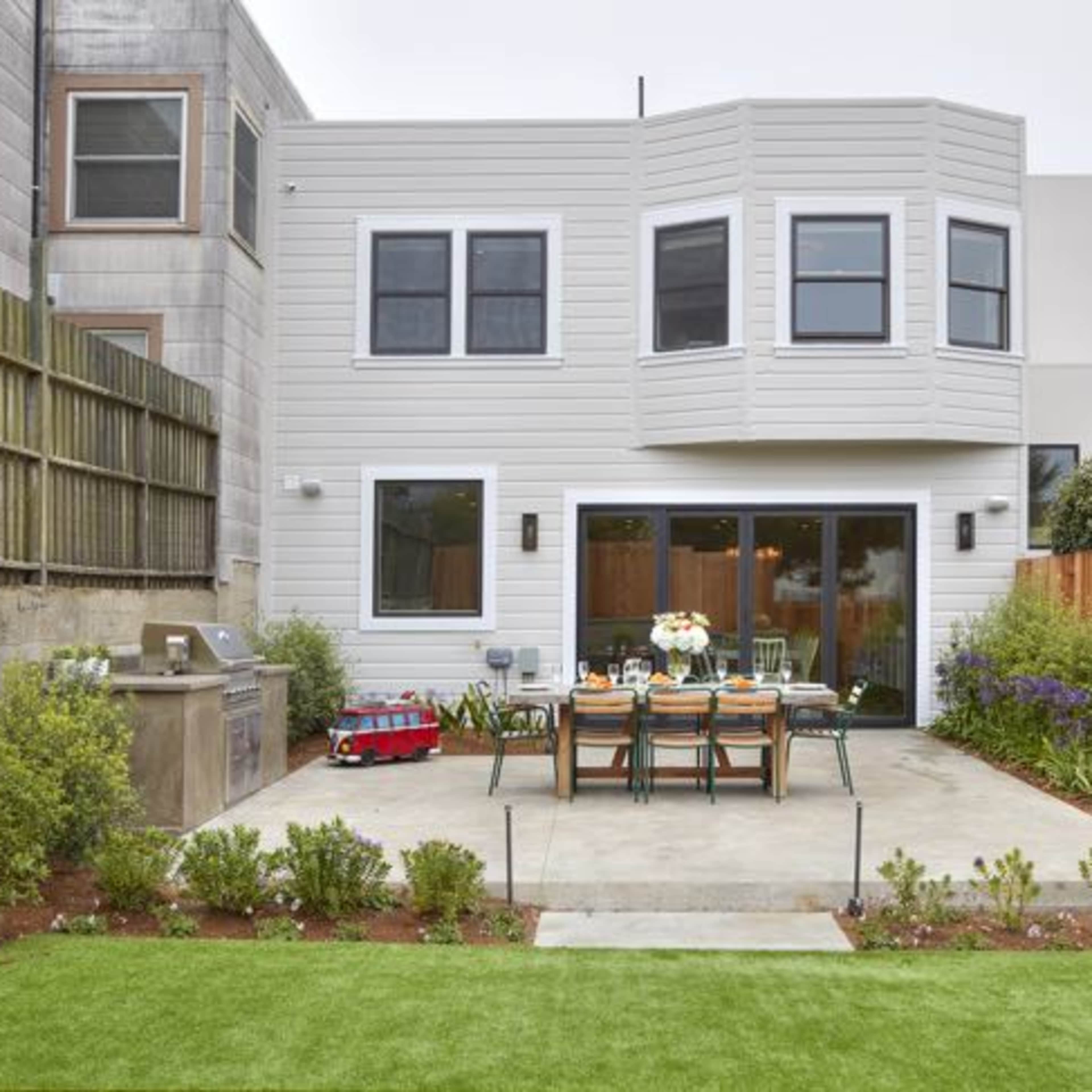 A modern backyard with a concrete patio, a dining table set for meals, a barbecue grill, and green landscaping.
