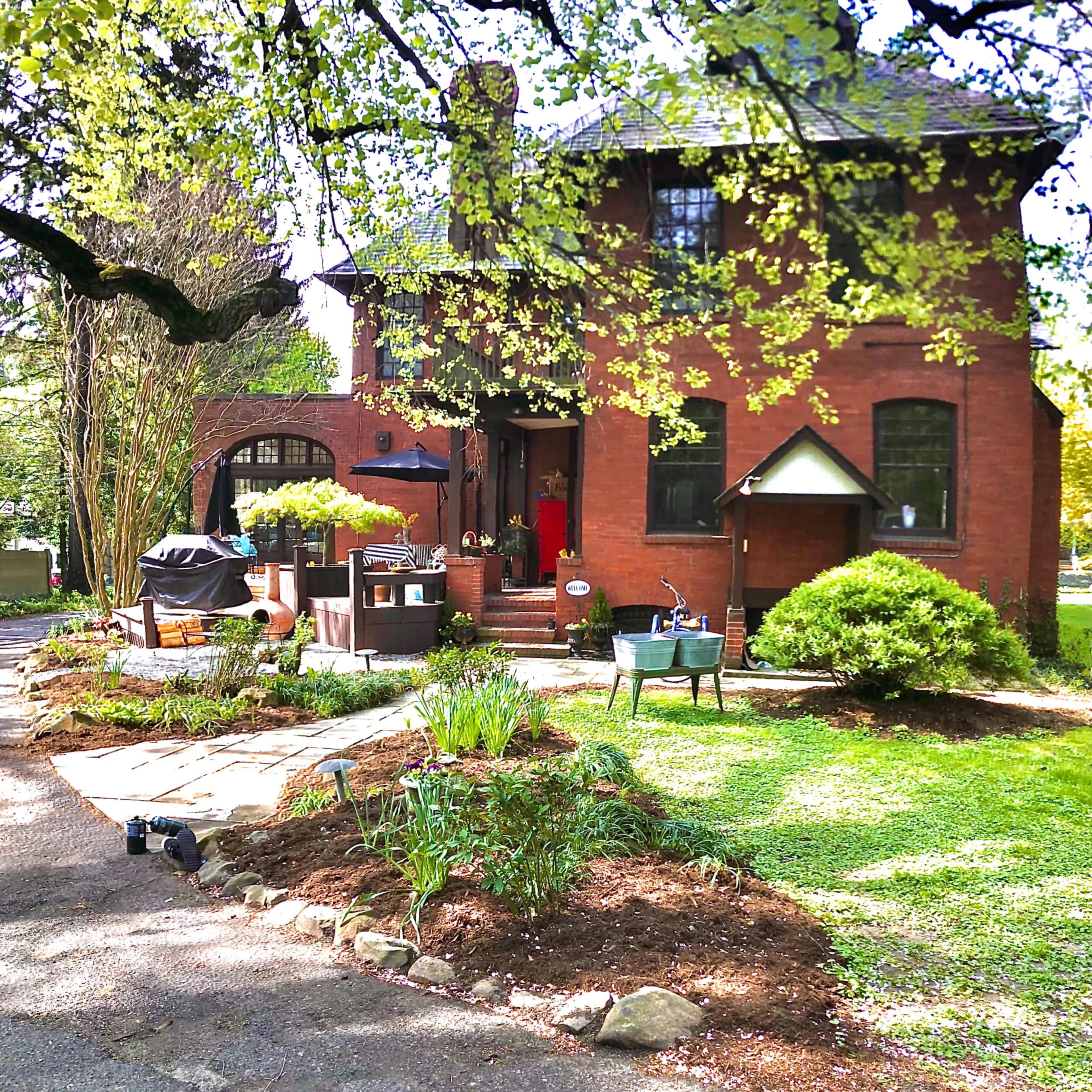 A brick house is surrounded by greenery and a landscaped yard with seating areas and a barbecue grill.
