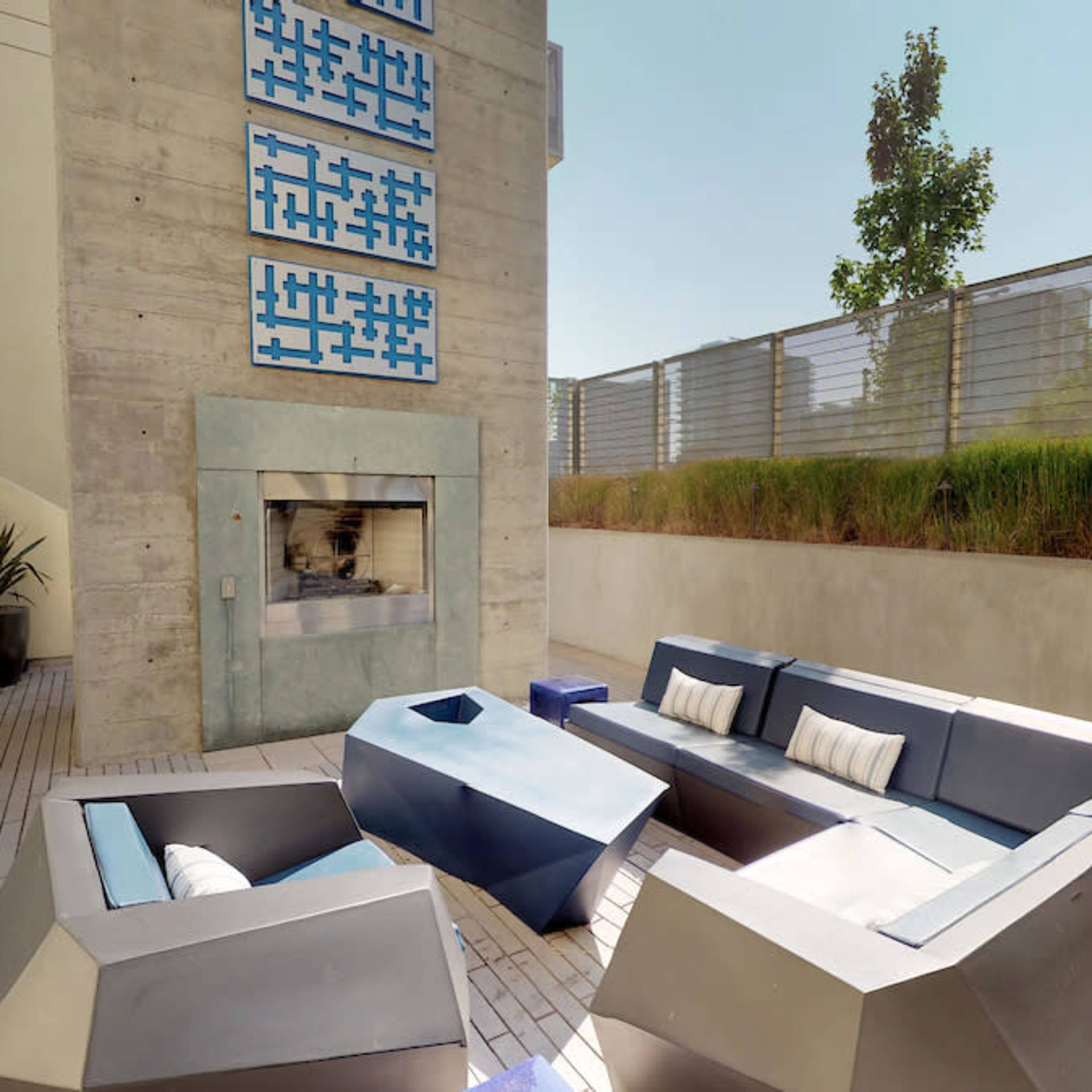 A modern outdoor seating area with a fireplace, featuring angular furniture and a minimalist design, set against a backdrop of greenery and a concrete wall.