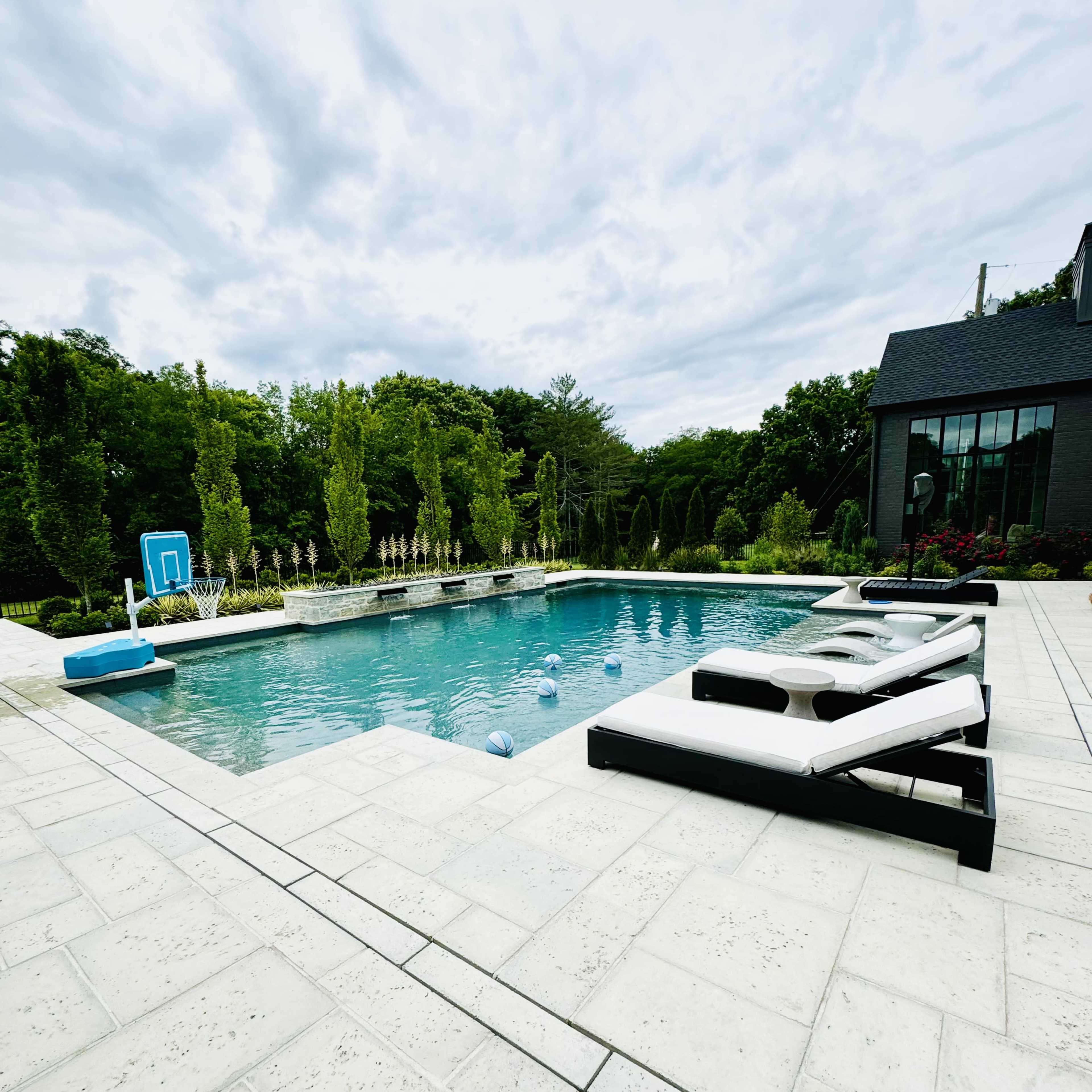 A rectangular swimming pool surrounded by a stone patio and lush green landscaping.