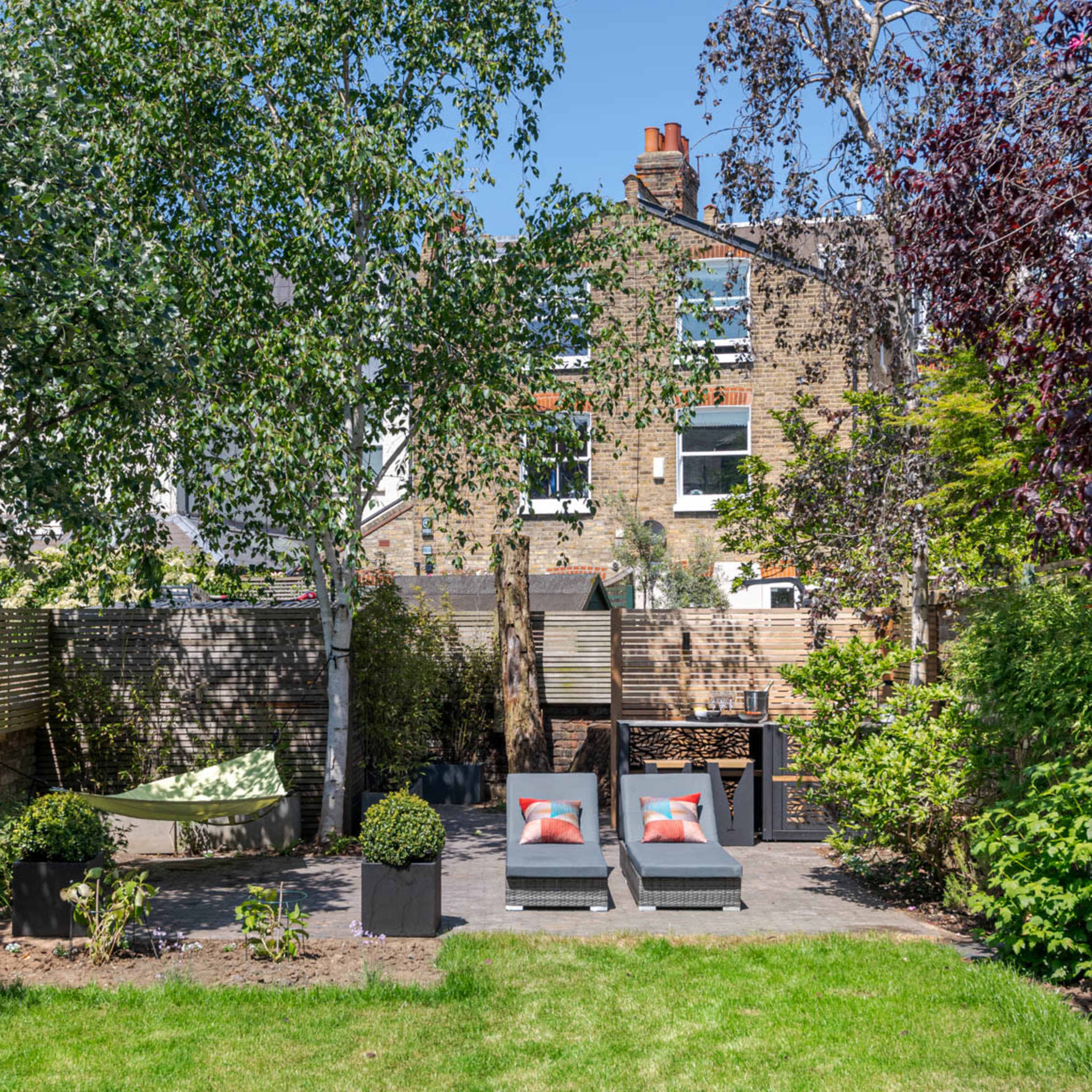 A landscaped backyard featuring two lounge chairs with cushions, surrounded by various plants and trees, against the backdrop of a multi-story building.