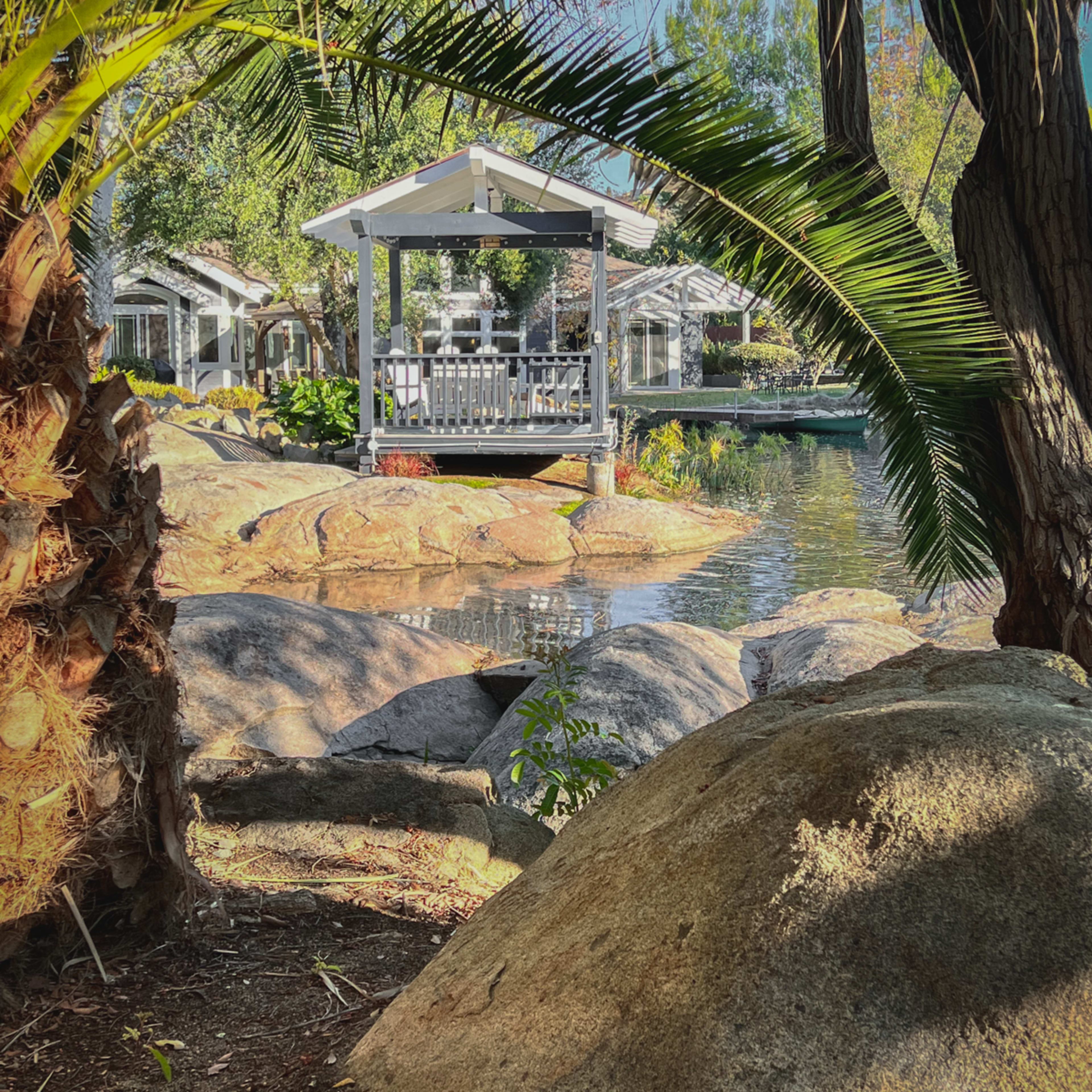A gazebo sits on the edge of a pond, surrounded by rocks and framed by palm fronds.