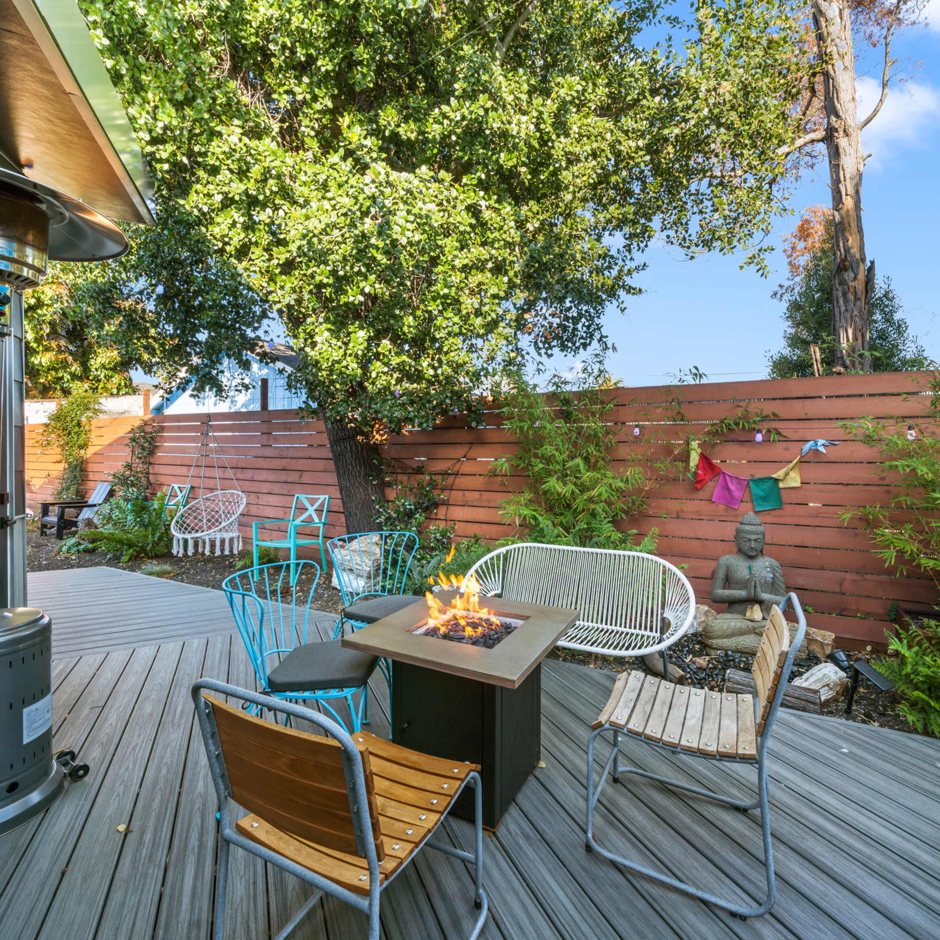 A wooden deck with a fire pit surrounded by several chairs and a statue, nestled among trees and a wooden fence.