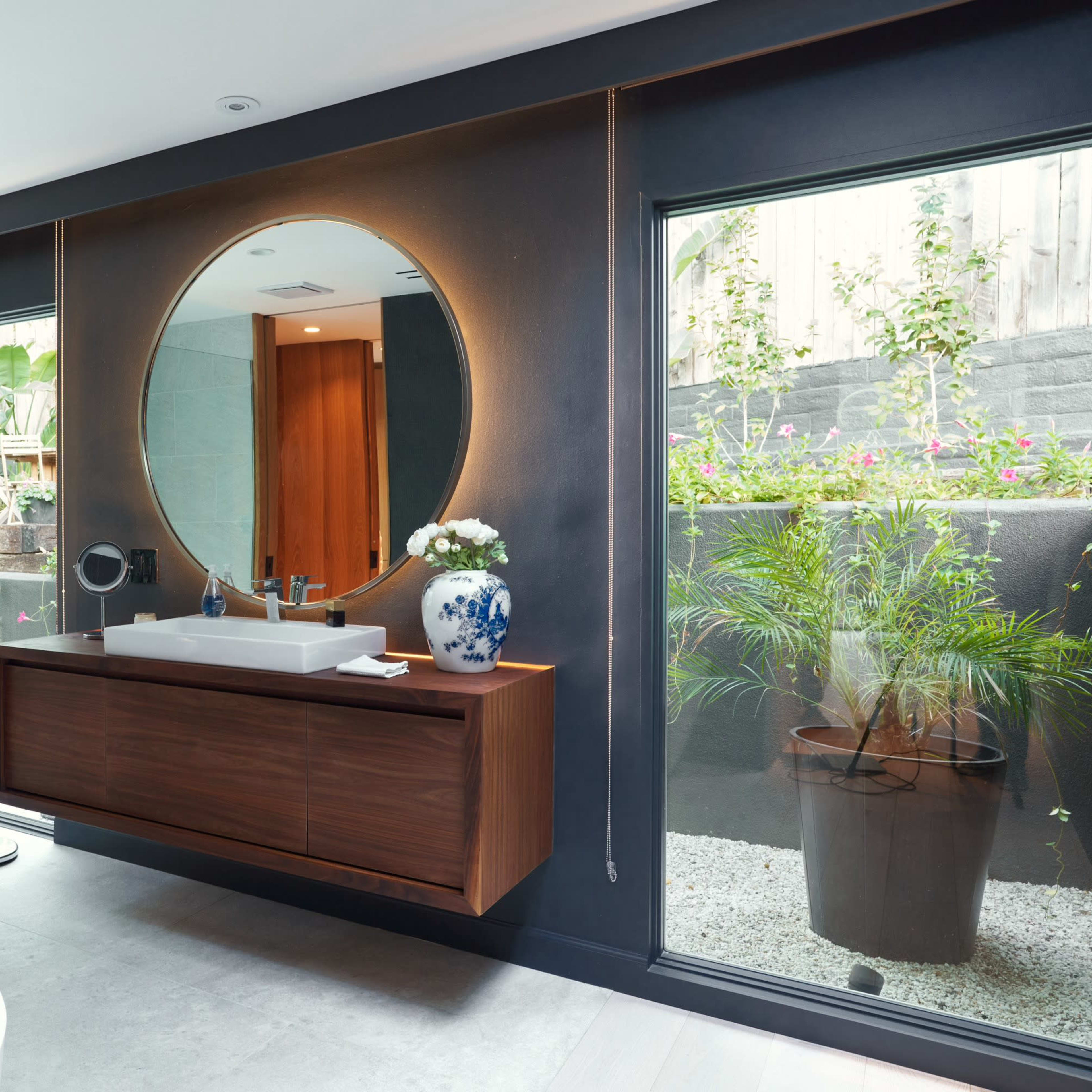 The bathroom features a round mirror above a wooden vanity, with large glass windows offering views of an outdoor garden area.