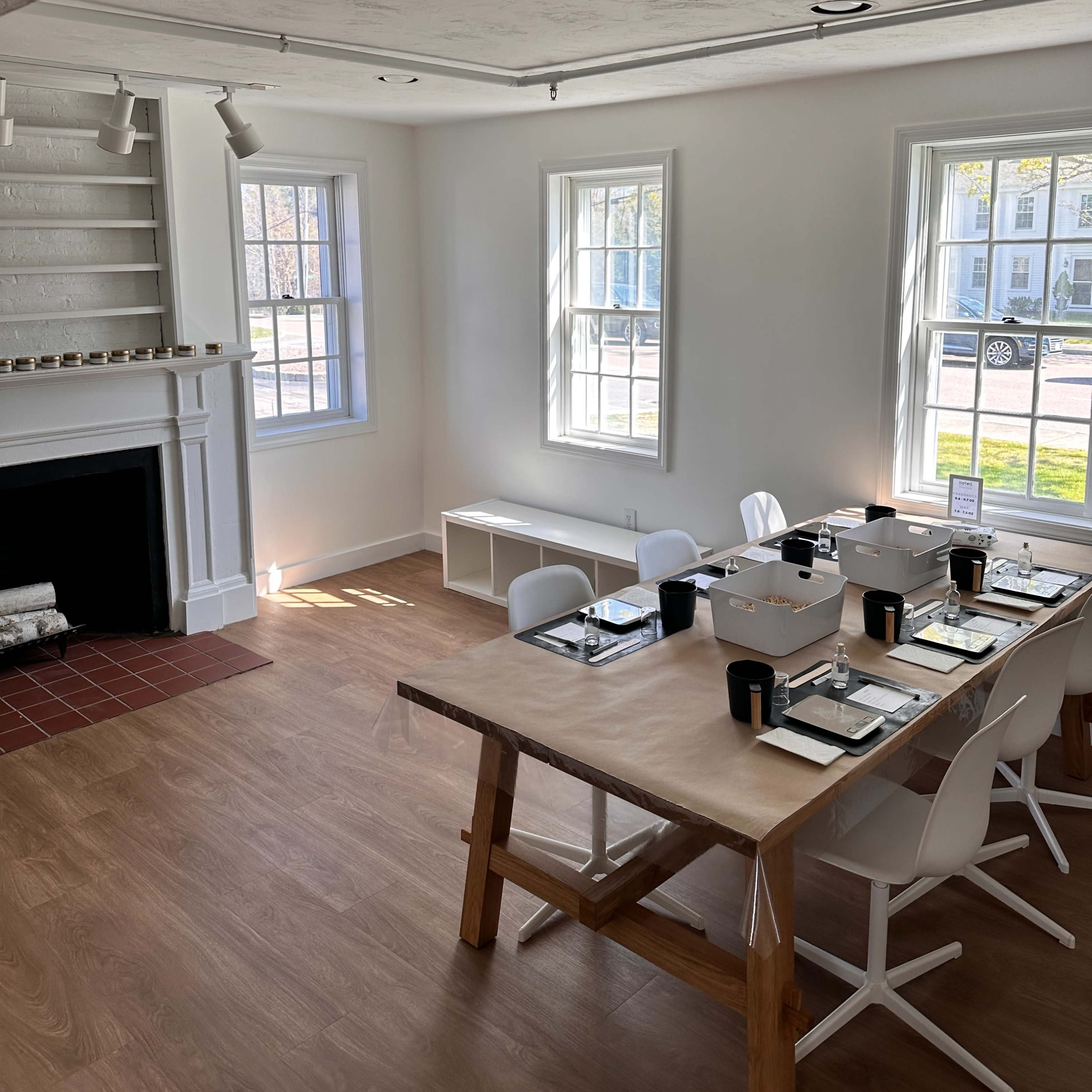 A bright, minimalist dining area with a large wooden table set for a meal, surrounded by white chairs and natural light pouring in through adjacent windows.