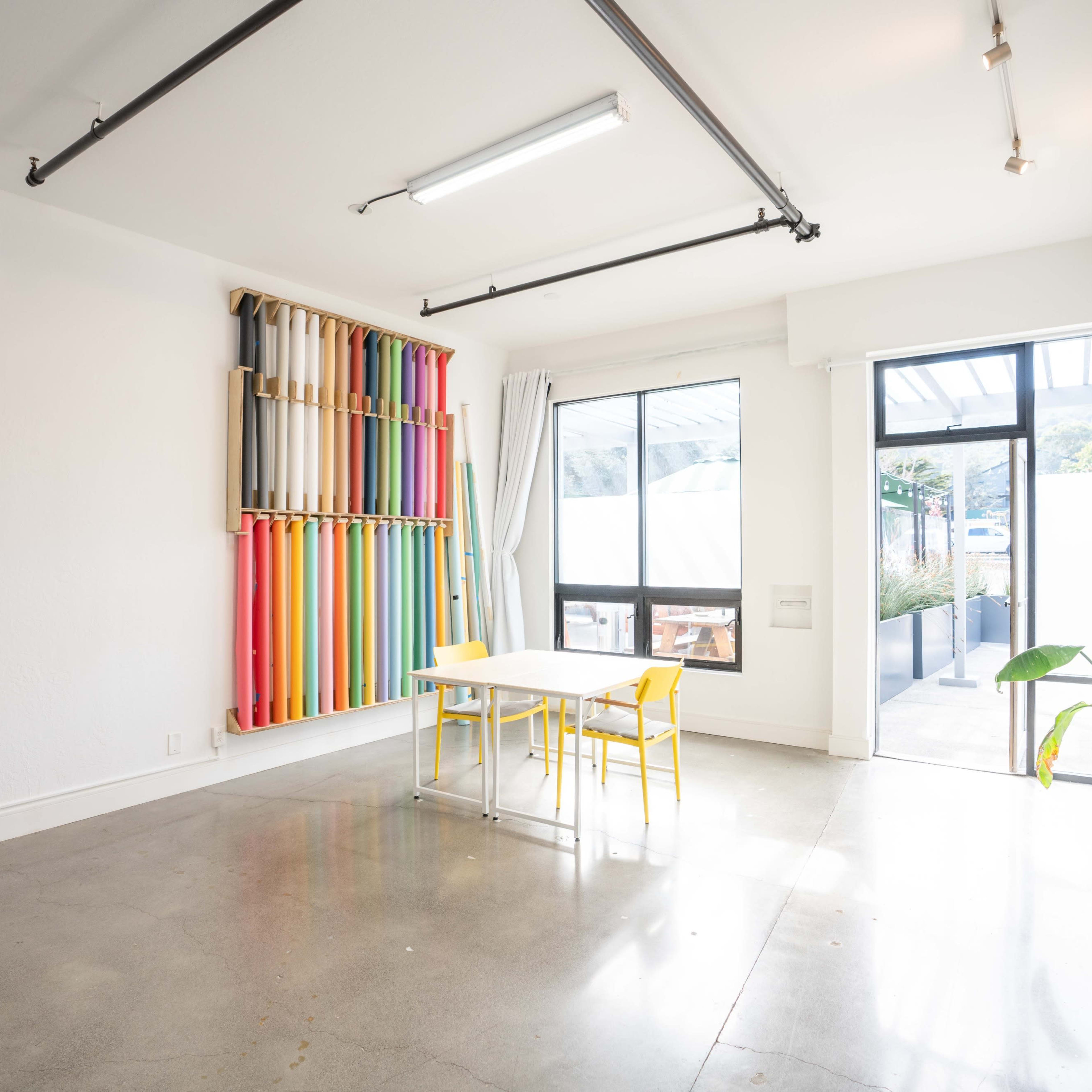 A minimalistic room with colorful fabric rolls on a wall, a white table, and two yellow chairs.